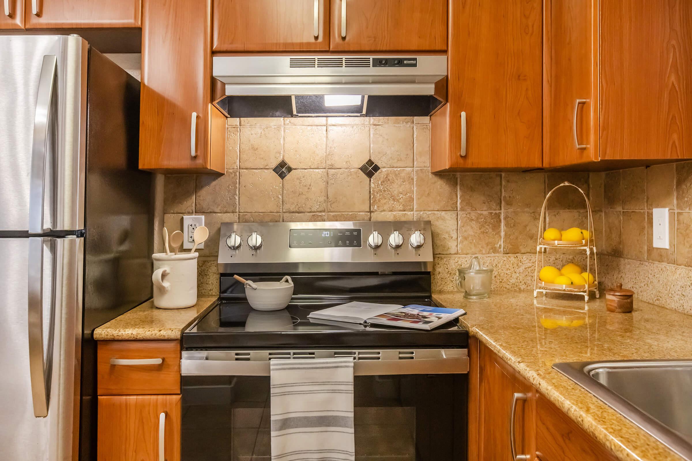 A modern kitchen featuring wooden cabinets, stainless steel appliances, and a tiled backsplash. The stove is centrally placed with a pot on it and a magazine nearby. A bowl of lemons sits in a basket on the counter, and there's a sink to the right, adding to the cozy, functional ambiance of the space.