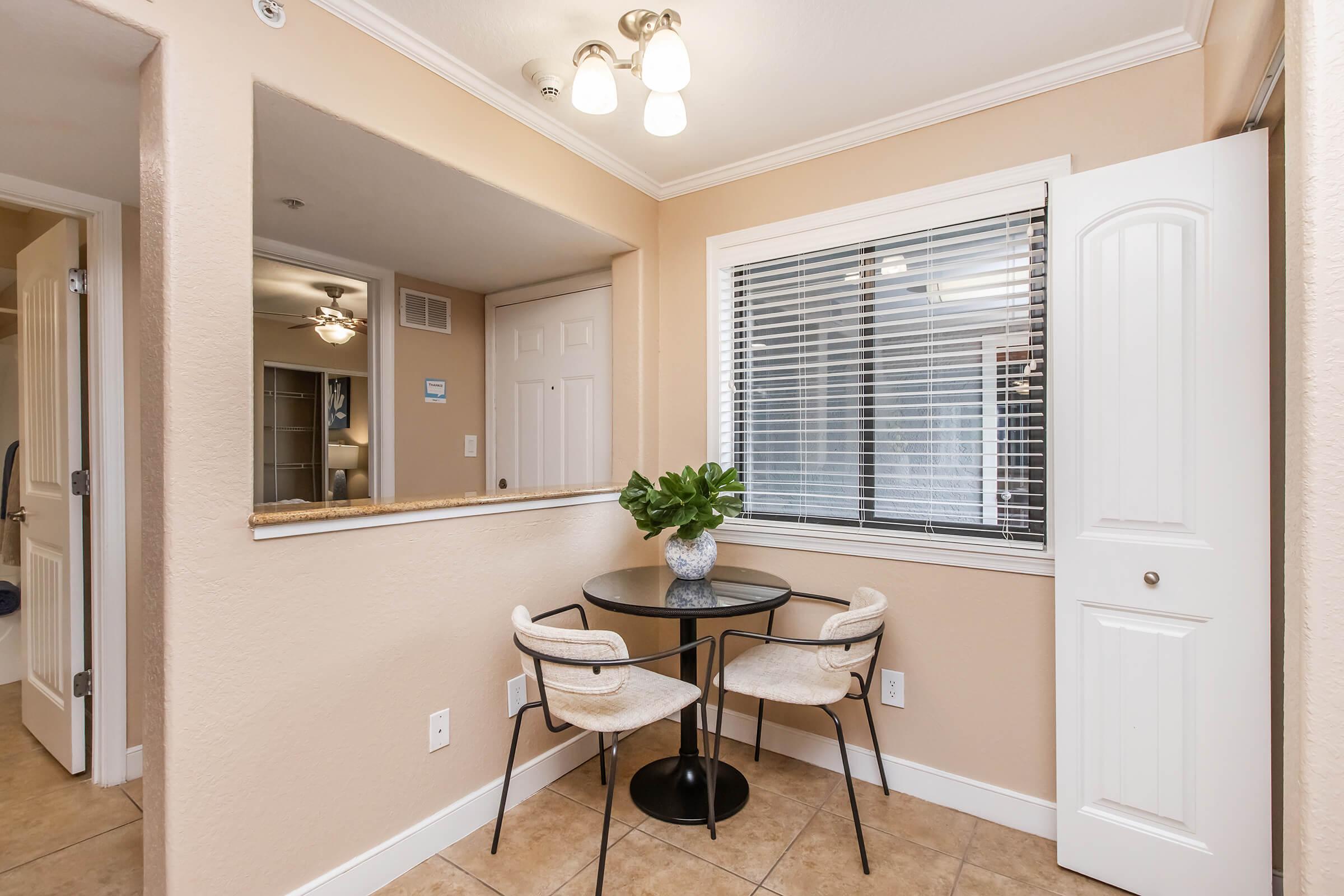 A small dining area features a round black table with two white chairs, set against a light beige wall. A potted plant sits on the table. A window with blinds allows natural light, and an open door leads to another room. The ceiling is adorned with a light fixture, enhancing the cozy atmosphere.