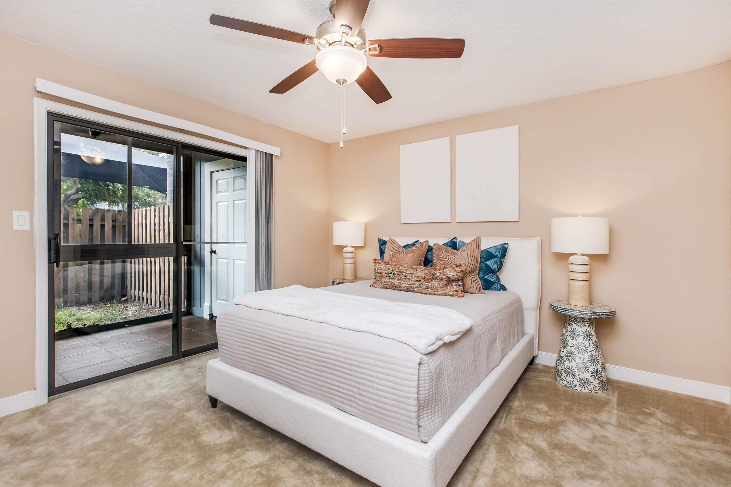 A stylish bedroom featuring a queen-sized bed with decorative pillows, a light-colored quilt, and two bedside lamps. A ceiling fan hangs above, and a sliding glass door leads to an outdoor area. The room has beige walls and a soft carpet, creating a cozy atmosphere.