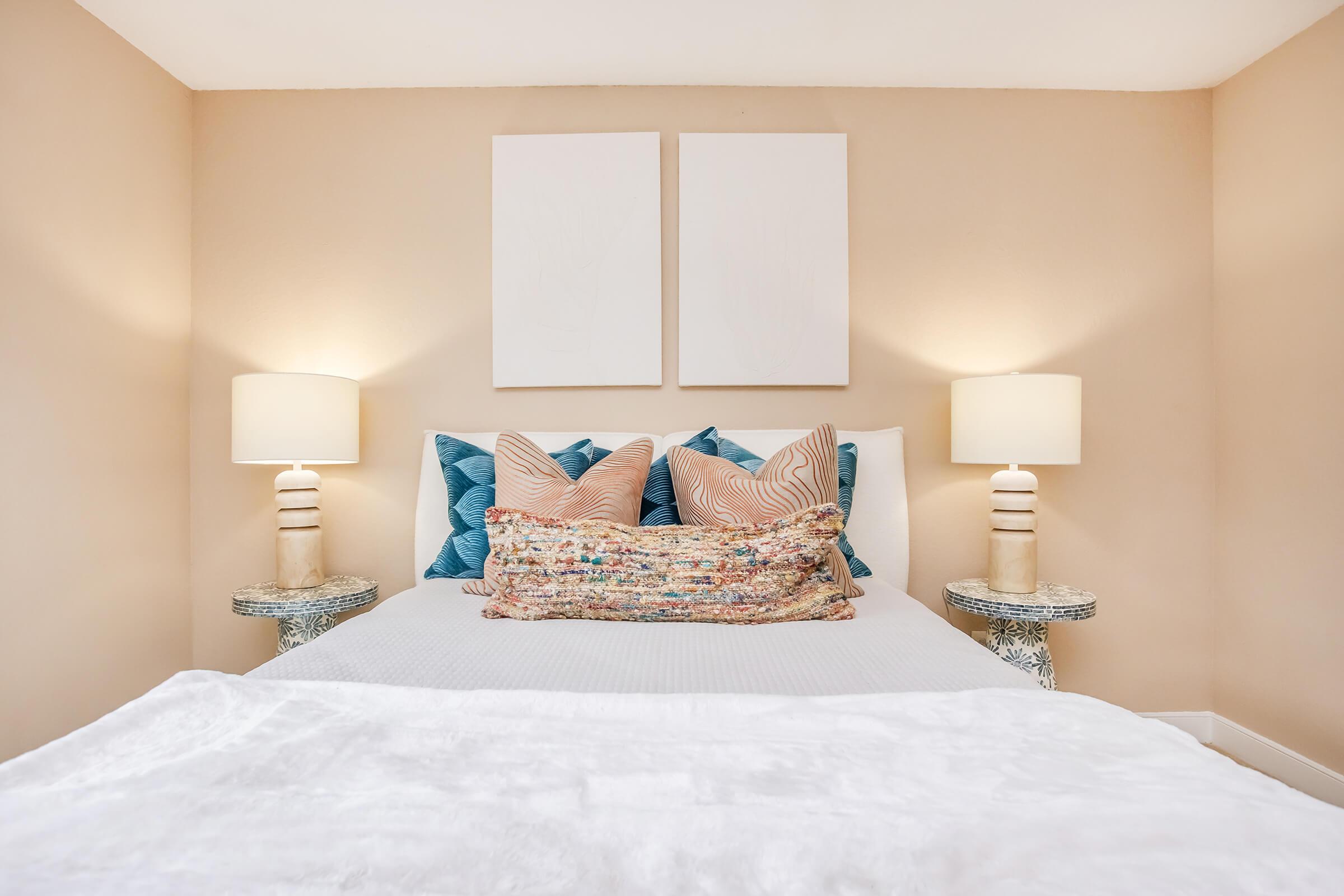 A cozy bedroom featuring a neatly made bed with decorative pillows in shades of blue and coral. There are two bedside tables with lamps providing soft lighting, and abstract art hangs on the wall above the headboard. The overall color scheme is soft and inviting, creating a relaxing atmosphere.