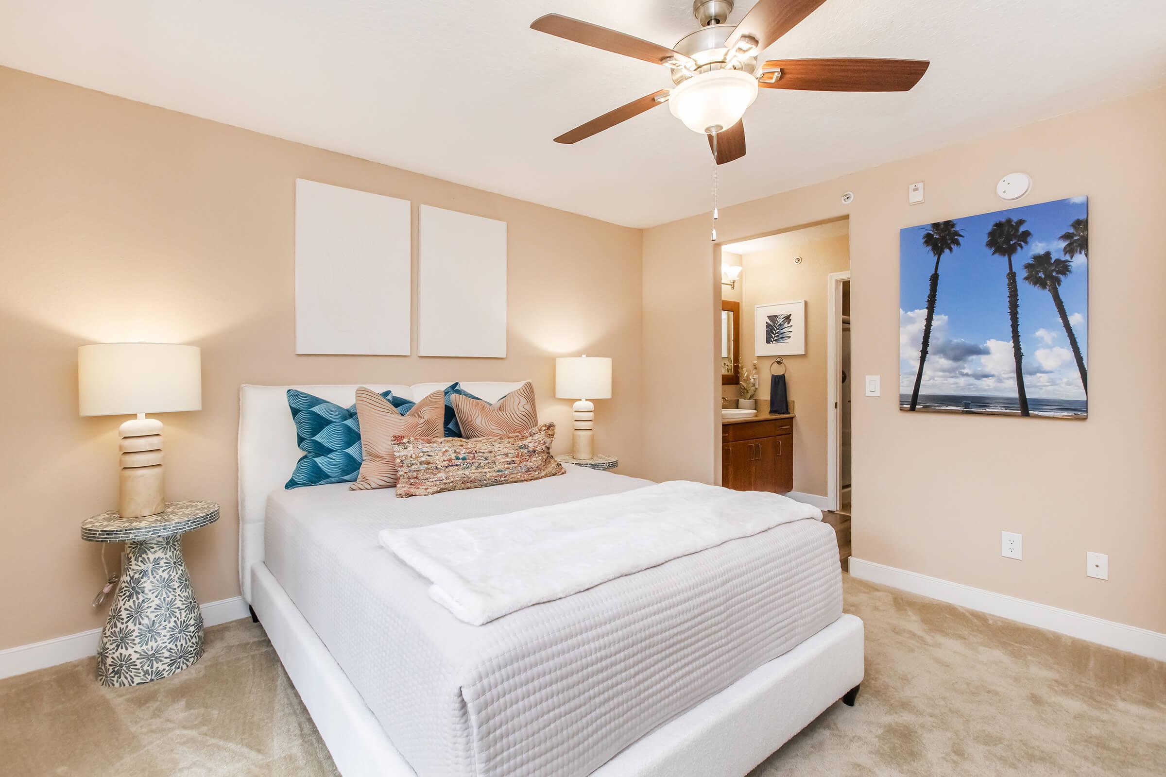 A cozy bedroom featuring a light-colored bed with decorative pillows, a stylish lamp on either side, and a ceiling fan. The walls are painted in a warm neutral tone, and there's a piece of artwork depicting palm trees and clouds. A bathroom door is visible in the background, adding to the room's inviting atmosphere.