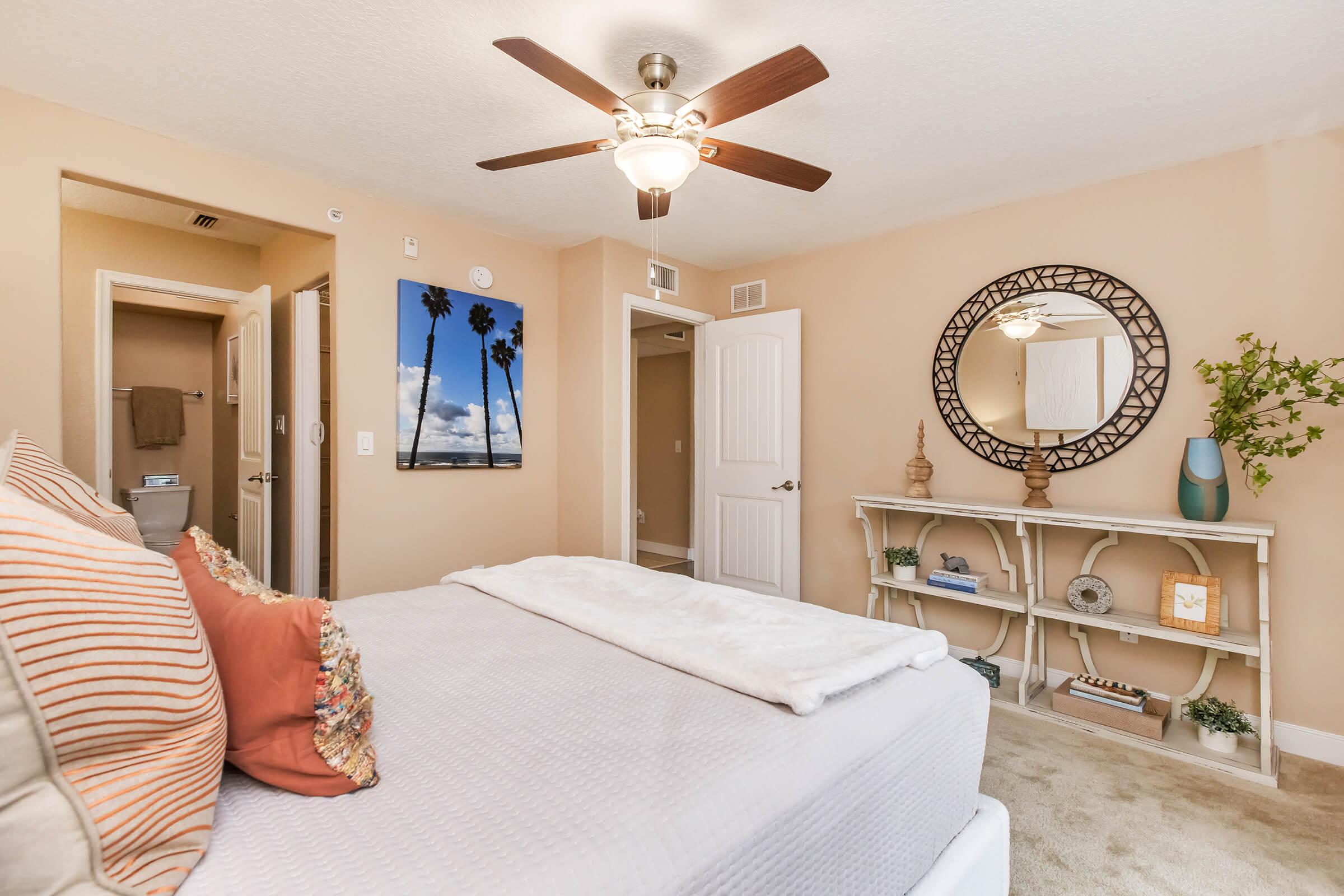 A bright and airy bedroom featuring a ceiling fan and a large mirror. The bed is dressed in a white comforter with decorative pillows in shades of orange. A wall-mounted photo of palm trees captures a coastal vibe. A small shelf holds decorative items, while a bathroom door is visible in the background.