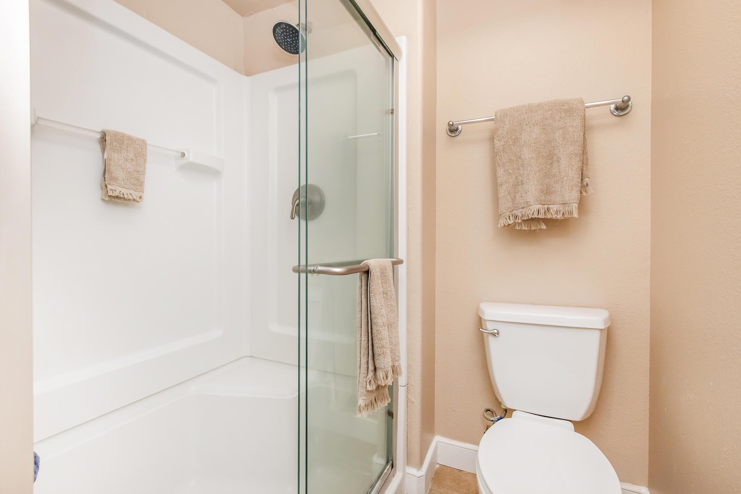 A modern bathroom featuring a clear glass shower enclosure with a white tub space, a white toilet, and two hanging beige towels on a rack. The walls are painted in a soft beige color, creating a warm and inviting atmosphere.