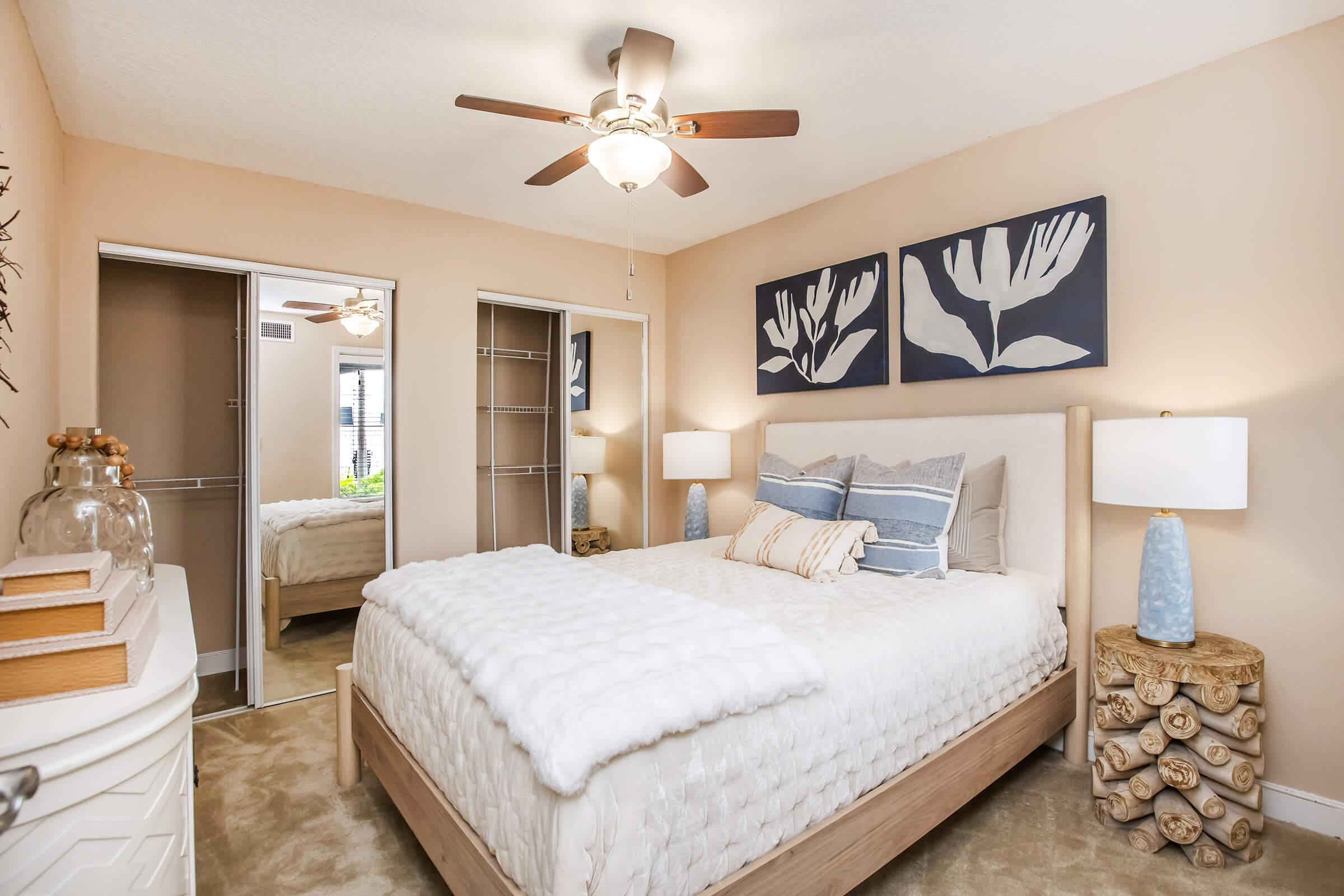 A cozy bedroom featuring a queen-sized bed with a light-colored comforter and decorative pillows. There's a wooden headboard, two bedside tables with lamps, and artwork on the walls depicting plants. A ceiling fan hangs above, and a closet with mirrored doors is visible in the background, adding to the room's spacious feel.