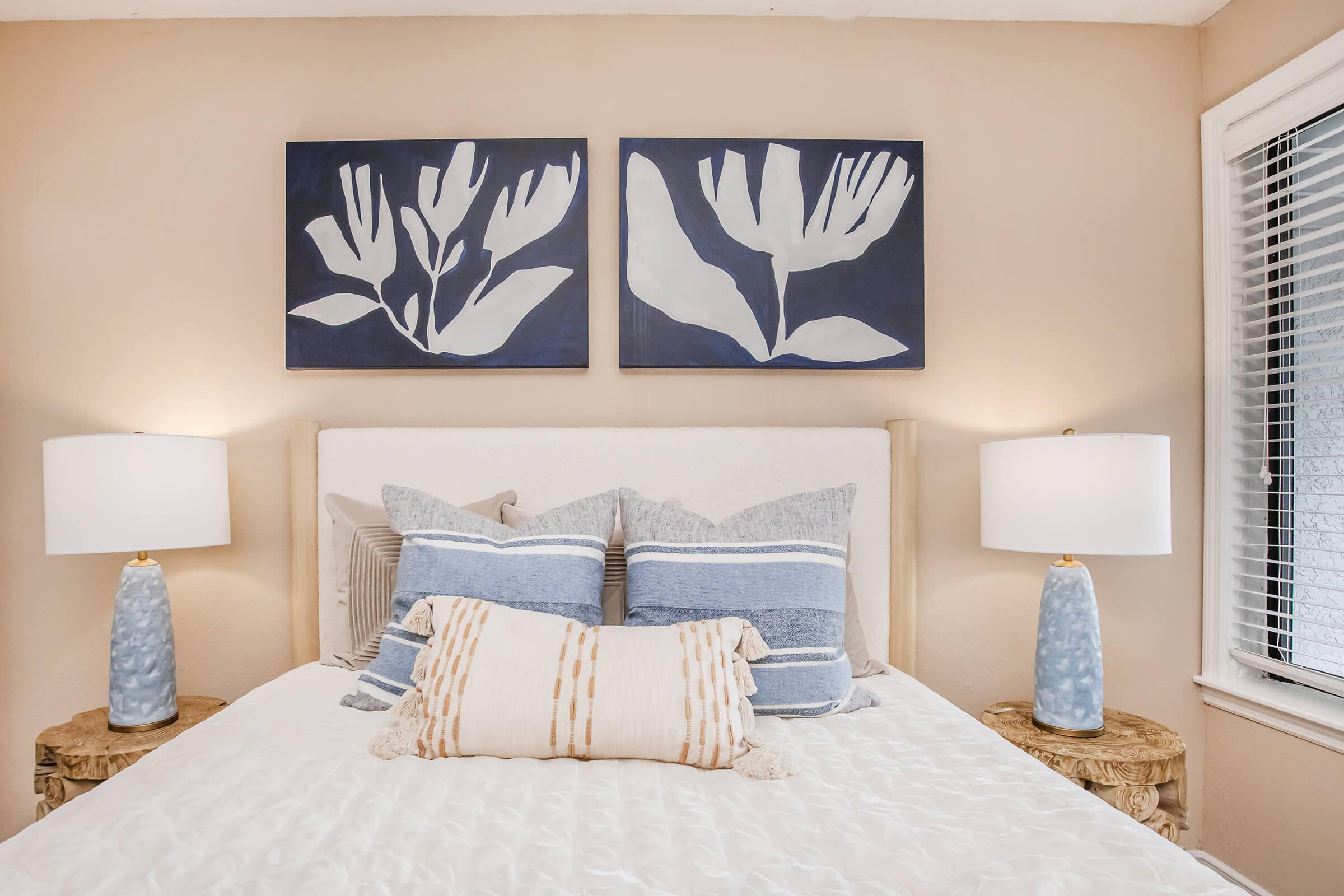 A cozy bedroom featuring a queen-size bed with white bedding and decorative pillows in shades of blue. On the wall above the bed, there are two abstract floral paintings. Each side of the bed has a lamp with a light blue base, and there are wooden nightstands with a natural finish. Natural light streams through a window.