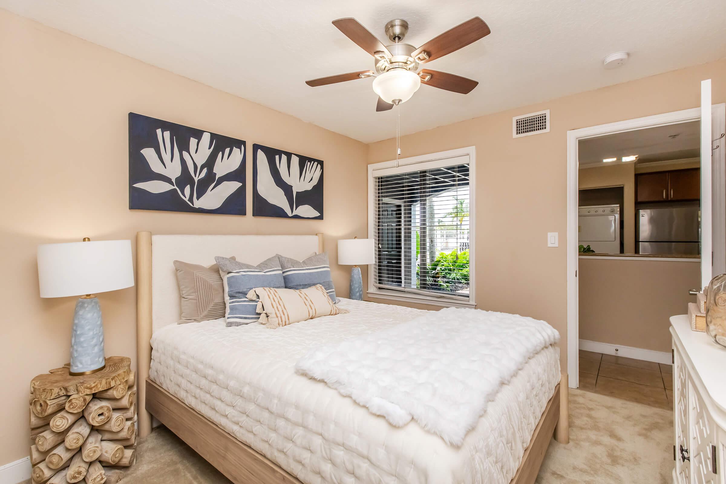 A cozy bedroom featuring a large bed with soft bedding, two decorative pillows, and a stylish headboard. There are two modern bedside lamps made of natural materials, and artwork with a floral theme on the wall. A window allows natural light to brighten the space, showcasing a view of greenery outside.