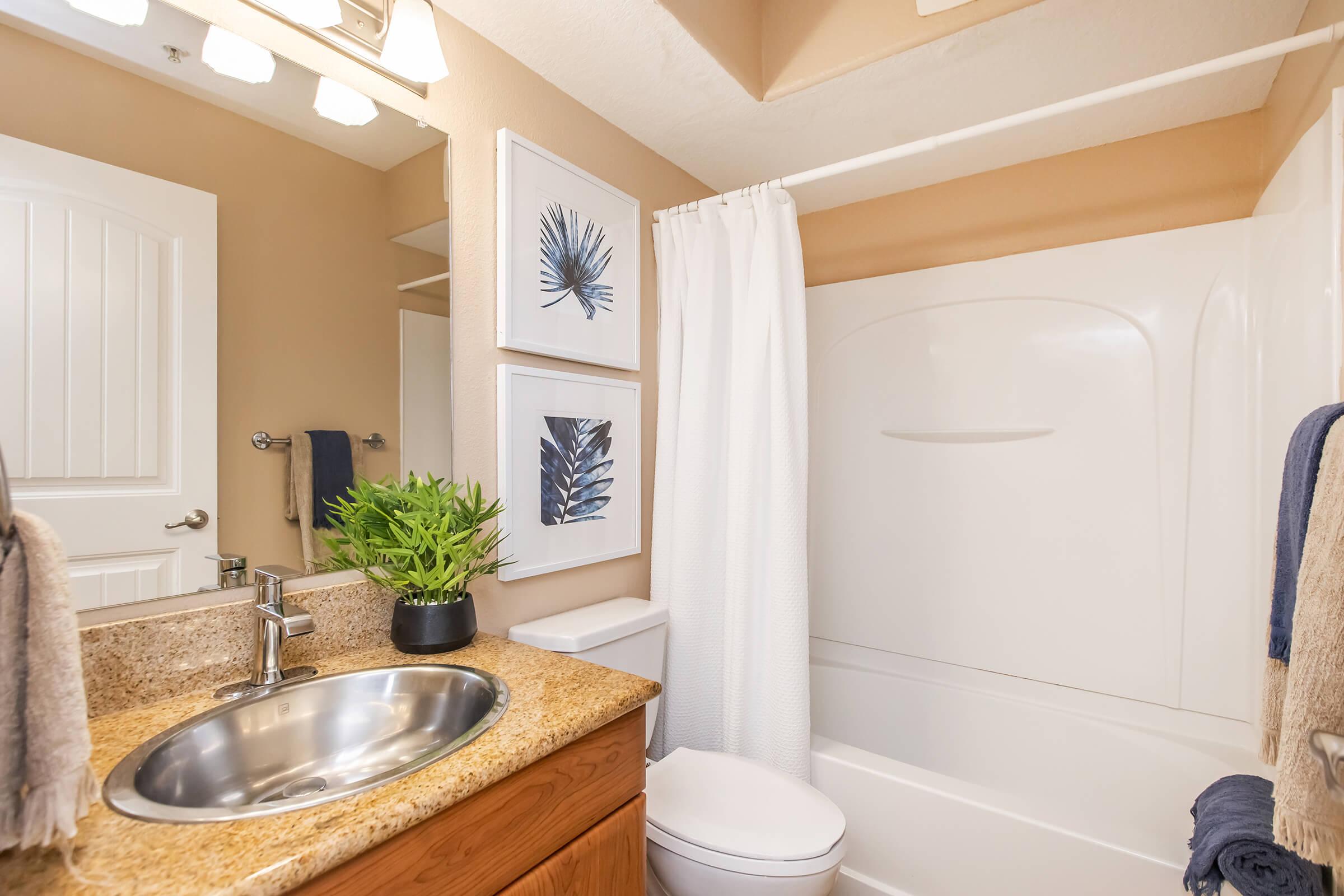 A well-designed bathroom featuring a white shower curtain, a sink with a stainless steel basin, wooden cabinetry, and a modern toilet. Wall art with botanical prints is displayed, and a potted plant adds a touch of greenery to the space. The color palette is neutral and inviting.
