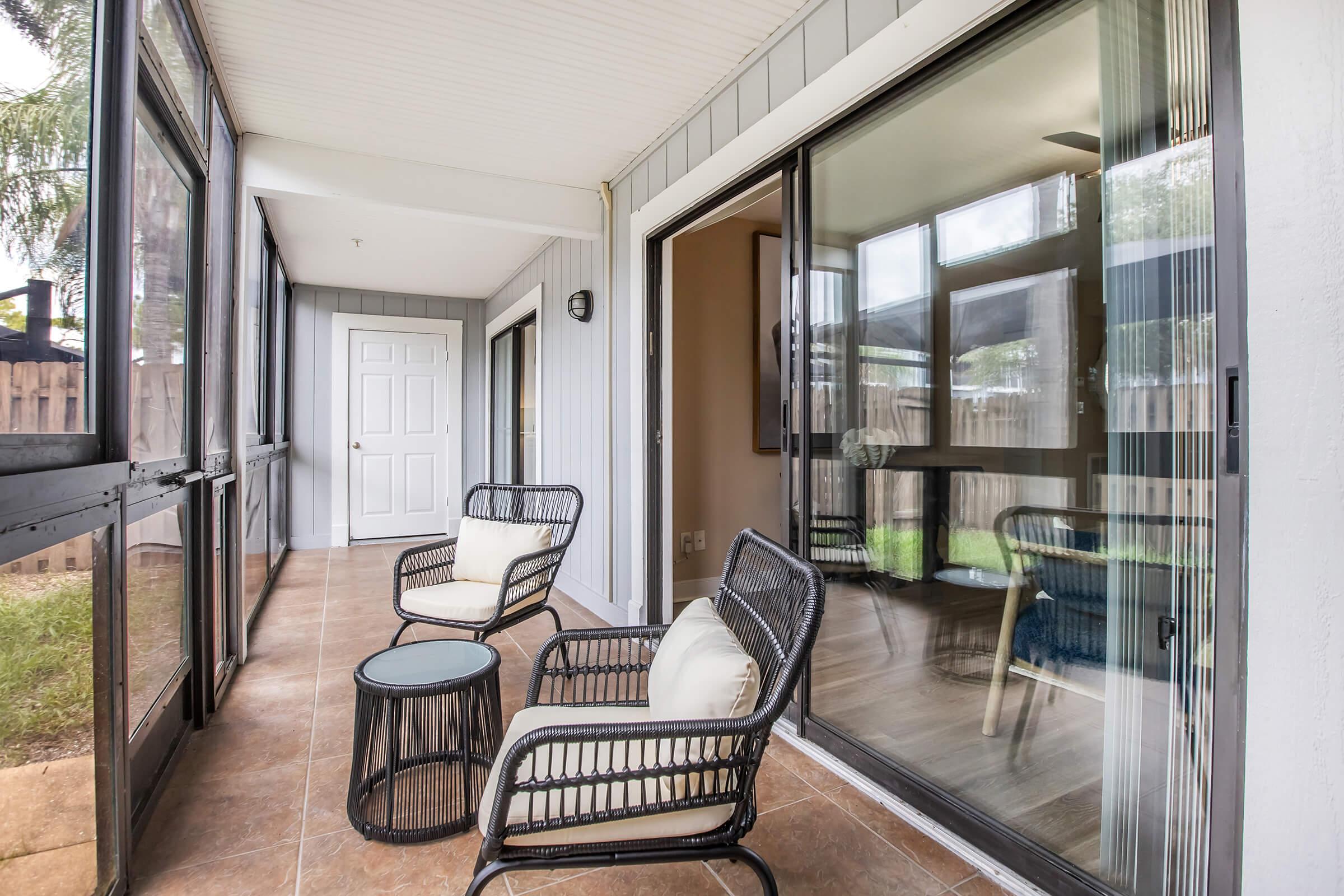 A cozy patio with two black wicker chairs and a round table, surrounded by glass doors. The space features neutral-colored walls and tiled flooring, creating a welcoming atmosphere. The view includes a green outdoor area through the transparent glass, enhancing natural light.