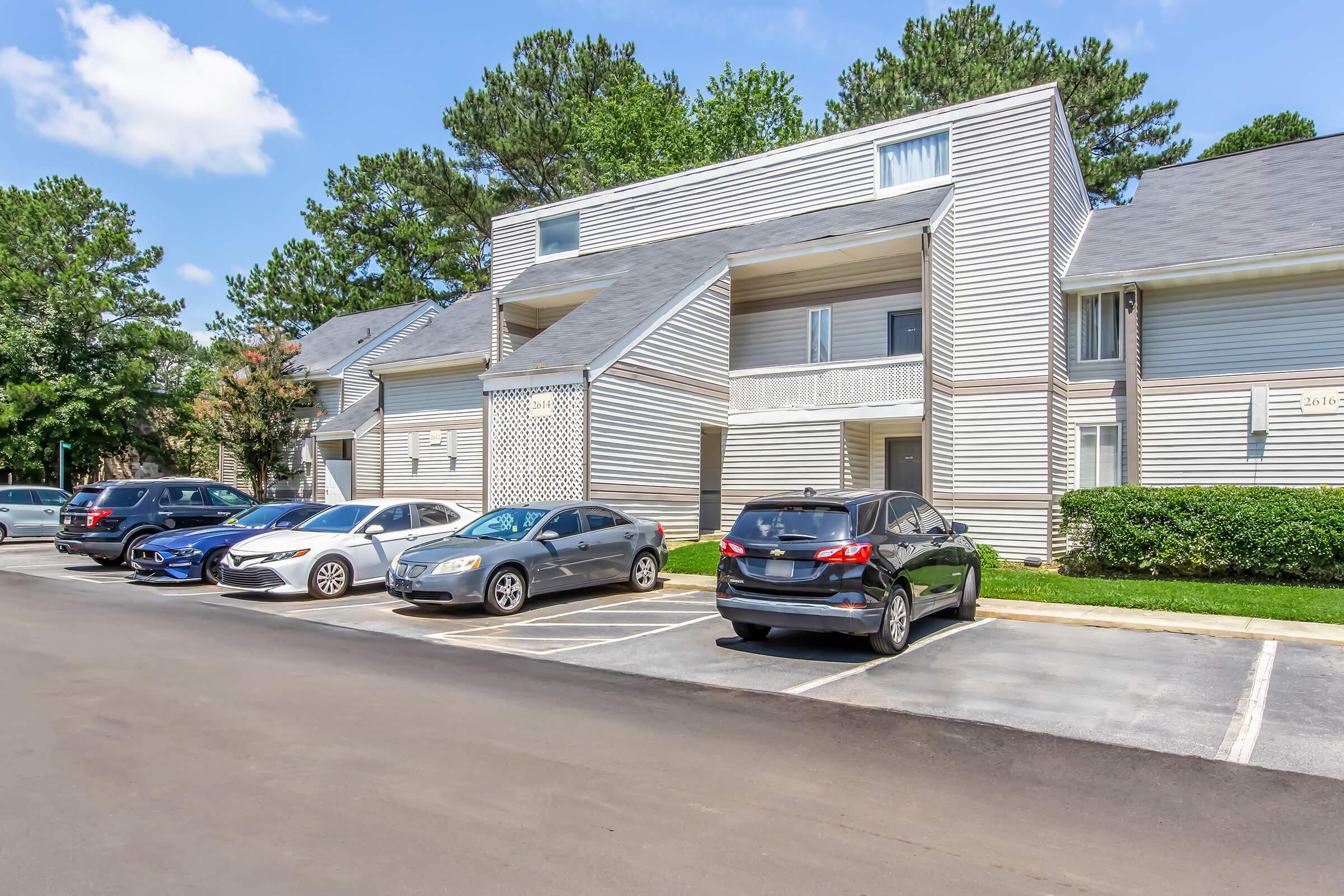 A row of modern apartment buildings with light wood siding, each featuring balconies. Several parked cars are lined up in front of the buildings on a paved parking lot. Lush green trees and blue sky with scattered clouds are visible in the background.