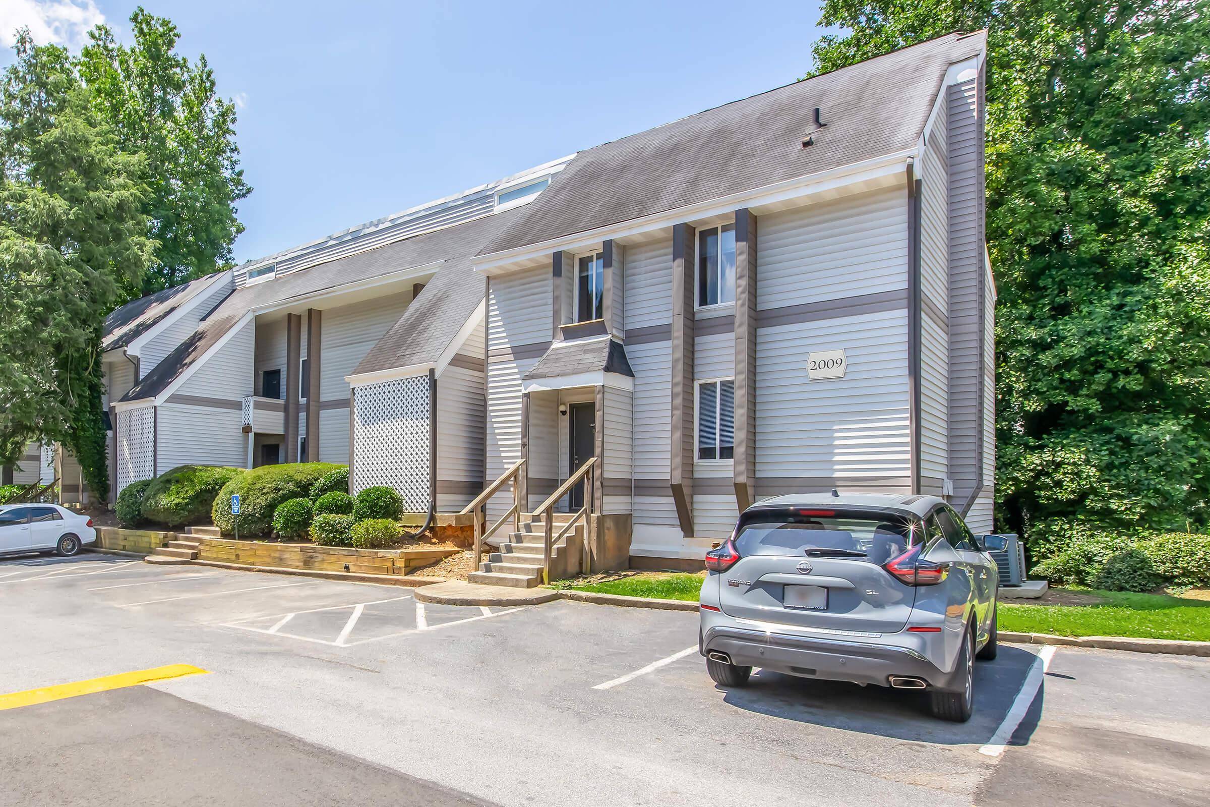 Two-story residential building with a light-colored exterior, featuring several staircases and balconies. In front, there is a parking area with a gray car parked in a space. Surrounding the building are manicured shrubs and trees, under a clear blue sky.