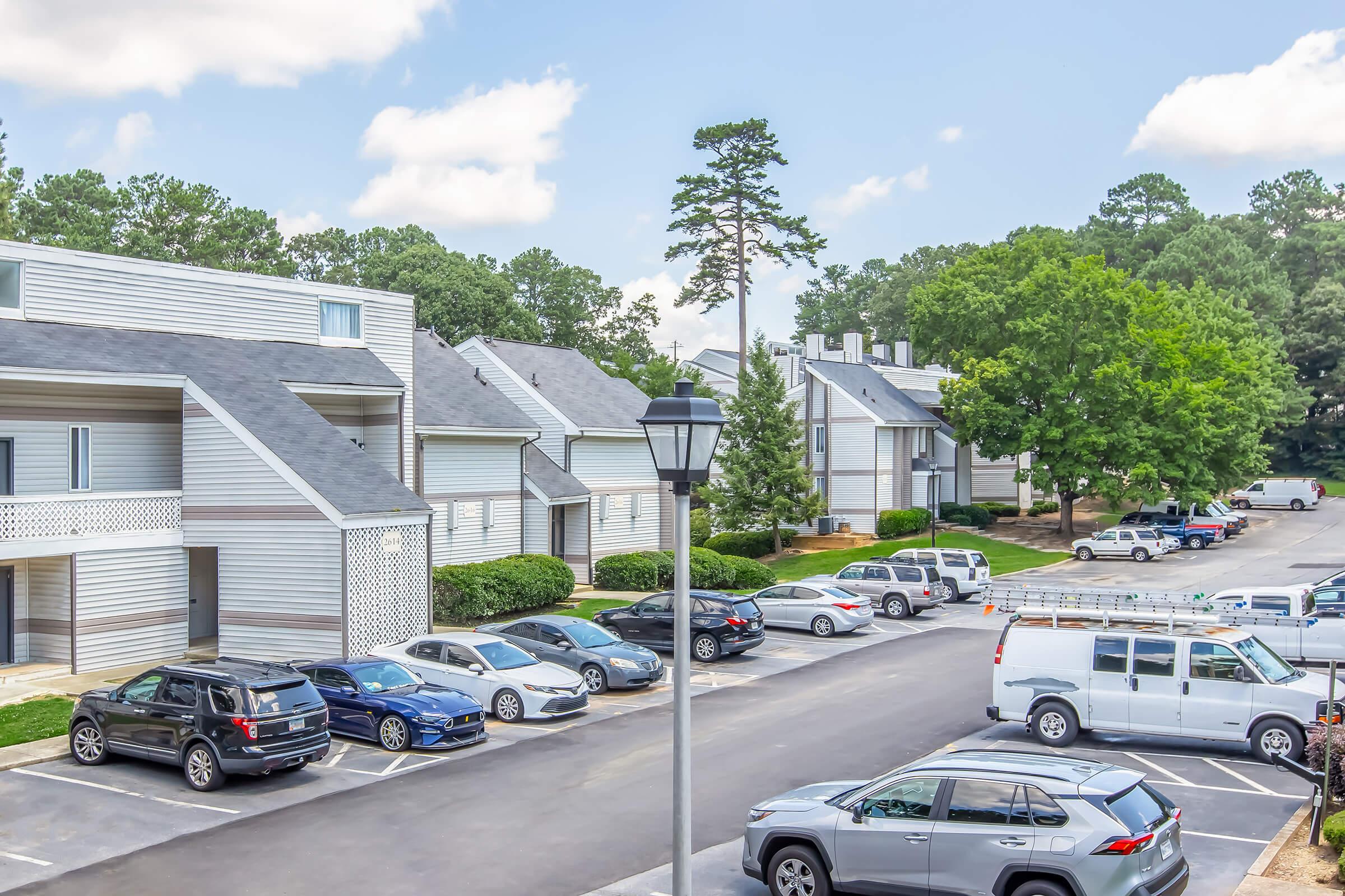 A residential parking area with several cars, surrounded by multi-unit apartment buildings and trees. The scene is bright and sunny, showing a development with green landscaping and a clear blue sky. A street lamp is visible in the foreground.