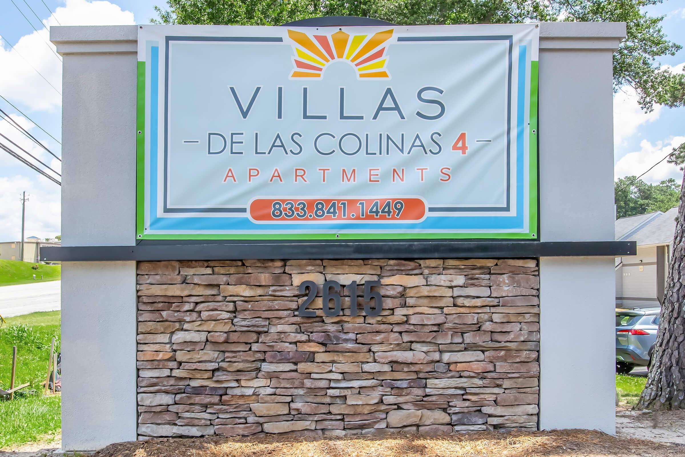 Signage for "Villas de las Colinas 4 Apartments" featuring a colorful logo with a sun design. The sign displays a contact number, 833.841.1449, and the address, 2015. The background shows a clear blue sky and greenery, highlighting the welcoming atmosphere of the apartment complex.