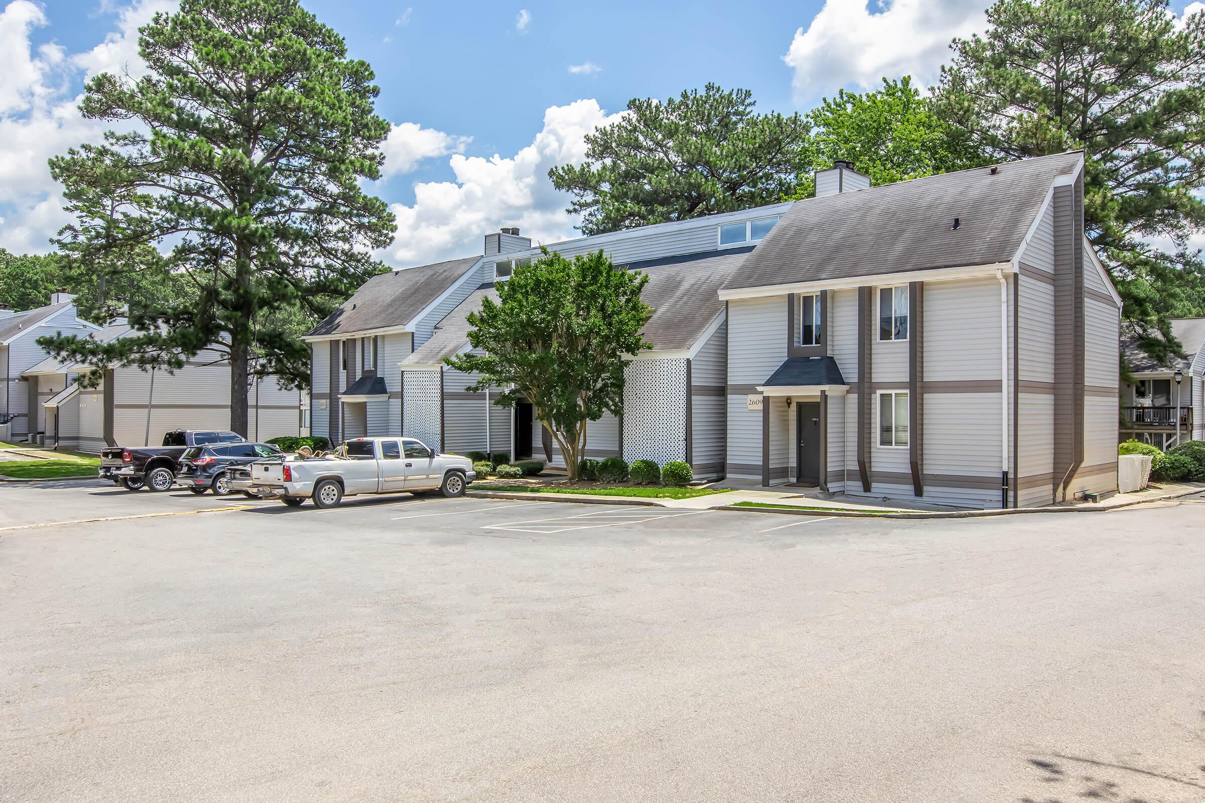 A residential complex featuring two-story apartment buildings with gray siding, surrounded by trees and green grass. A parking lot in the foreground includes several vehicles. The scene is set under a blue sky dotted with fluffy white clouds.