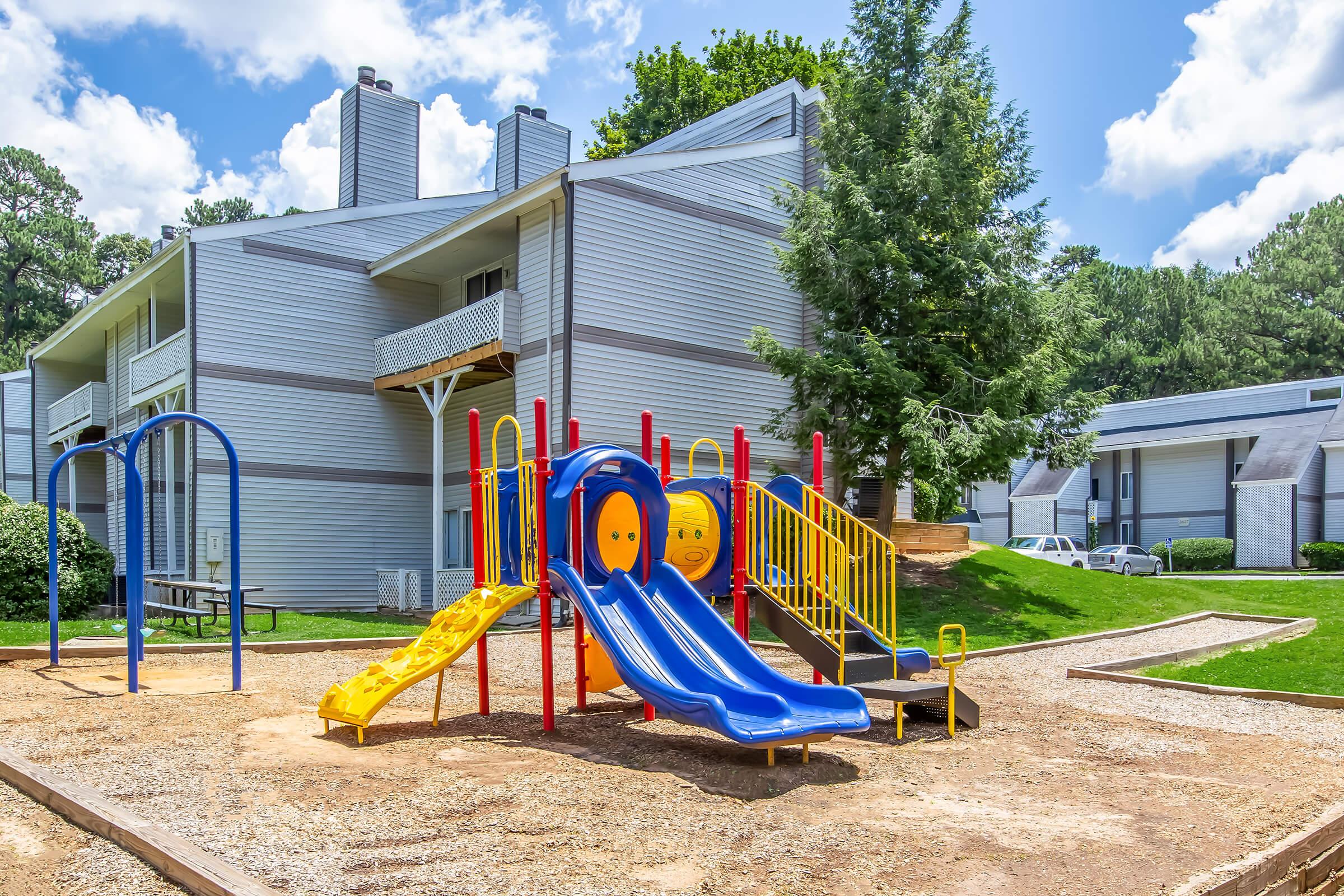 Colorful playground featuring yellow and blue slides, climbing equipment, and swings, set on a sandy area. In the background, there are two apartment buildings surrounded by green grass and trees under a bright blue sky with fluffy white clouds.