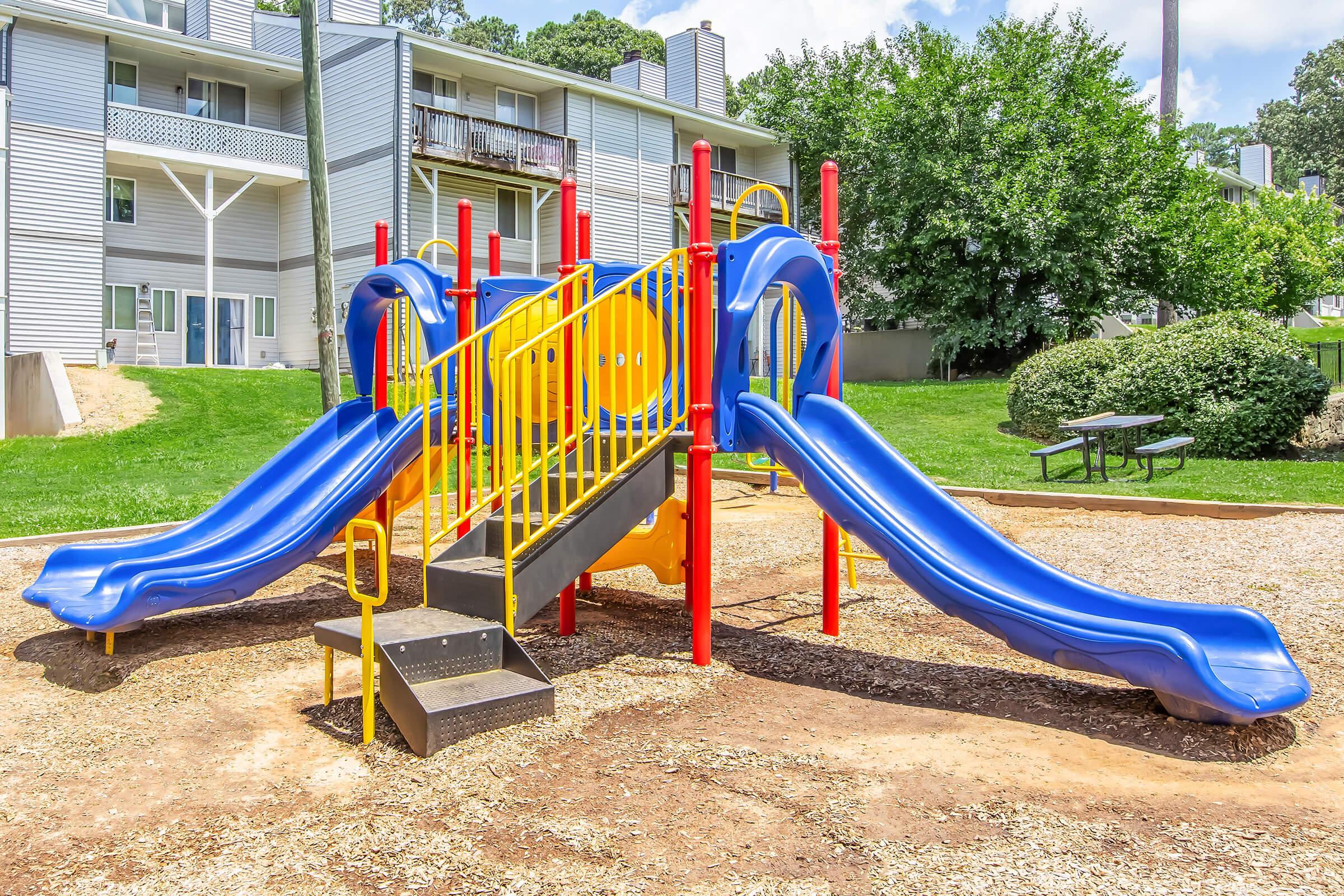 A colorful playground structure featuring two blue slides, a climbing area, and safety railings, situated on a sandy surface. In the background, there are residential buildings and green foliage, creating a pleasant outdoor play environment.