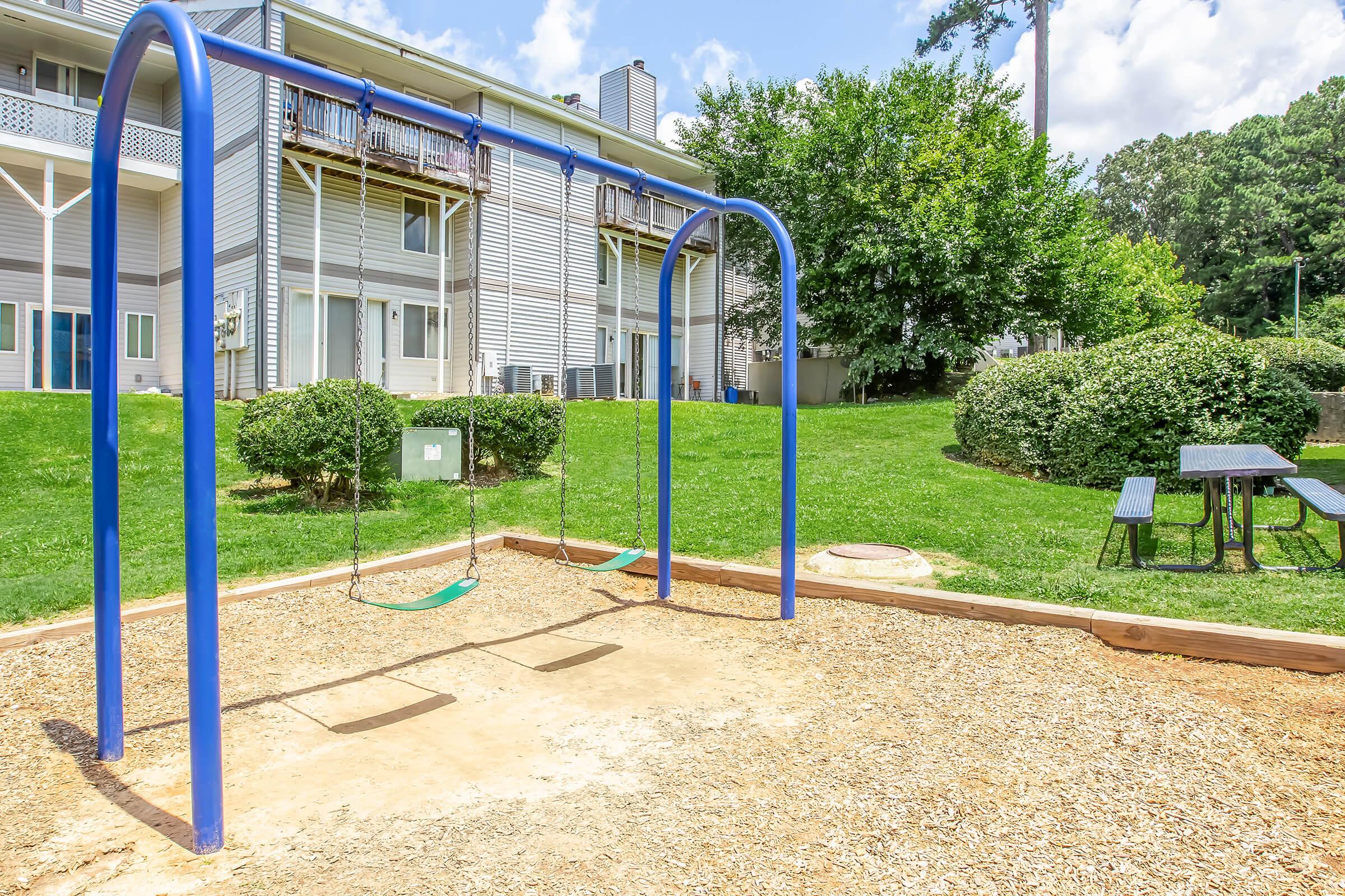 A playground featuring a set of blue swings on a wooden frame, surrounded by a grassy area and shrubs. In the background, apartment buildings are visible. The scene is bright and sunny, with a picnic table nearby, creating a family-friendly environment.