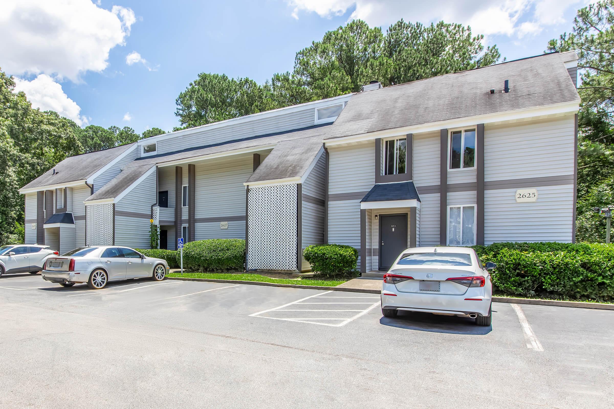Exterior view of a two-story apartment building surrounded by greenery, with a blue sky and clouds above. The building features a modern facade, with windows on both floors. Several parked cars are visible in the foreground, along with clearly marked parking spaces.