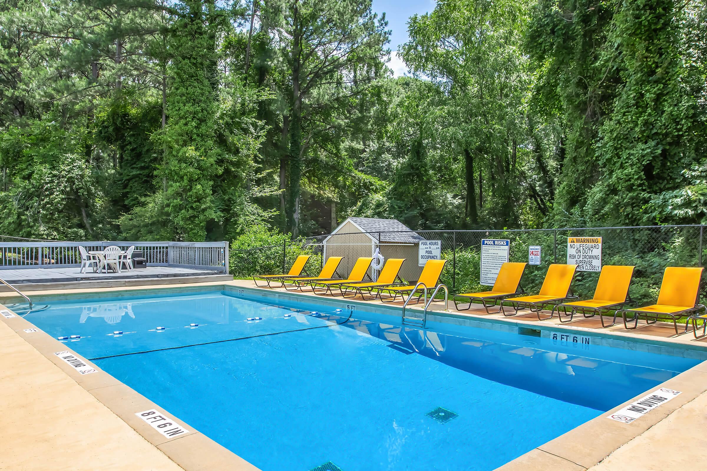 A clear swimming pool surrounded by lush greenery. Yellow lounge chairs are lined up by the pool's edge, and there's a small wooden deck with a table and chairs nearby. Signs are displayed around the area, providing information about pool rules and safety.