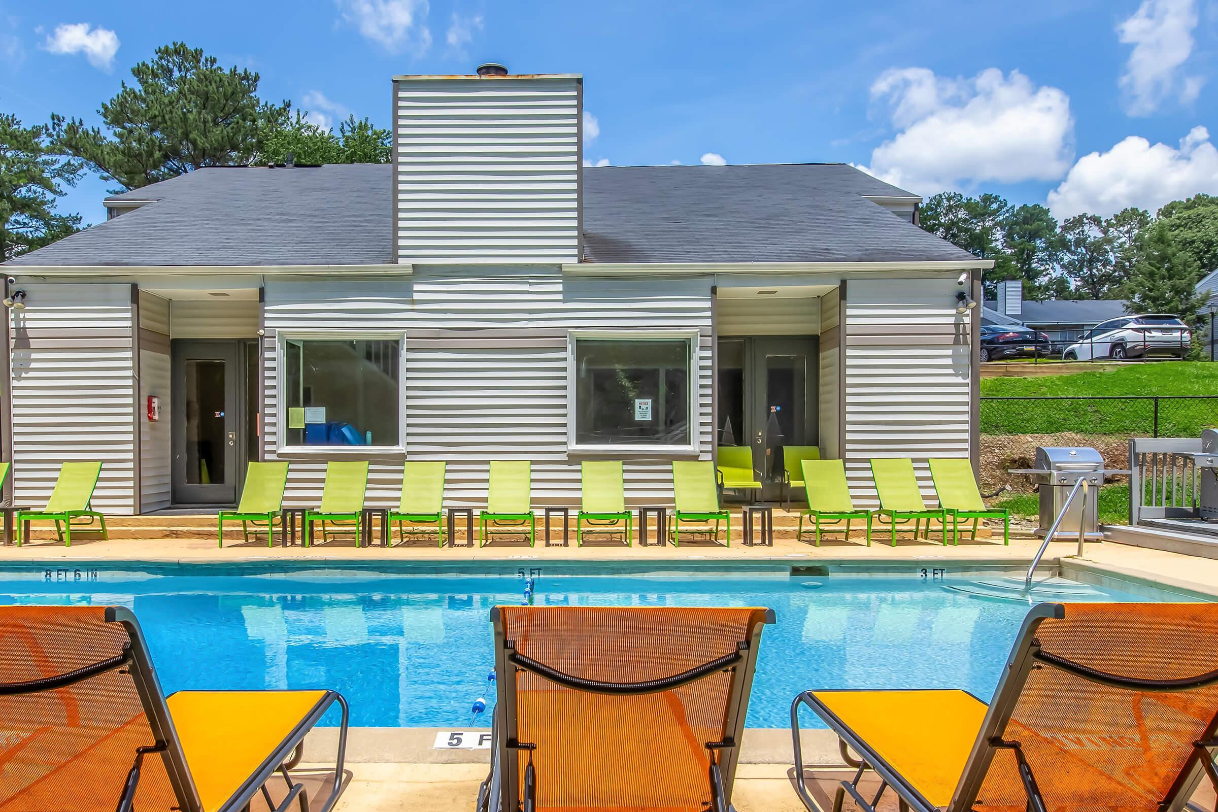 A serene swimming pool in a residential area, featuring bright yellow lounge chairs lined up on the deck. Behind the pool, a modern building with large windows and a chimney. Lush greenery surrounds the property under a clear blue sky with fluffy white clouds.