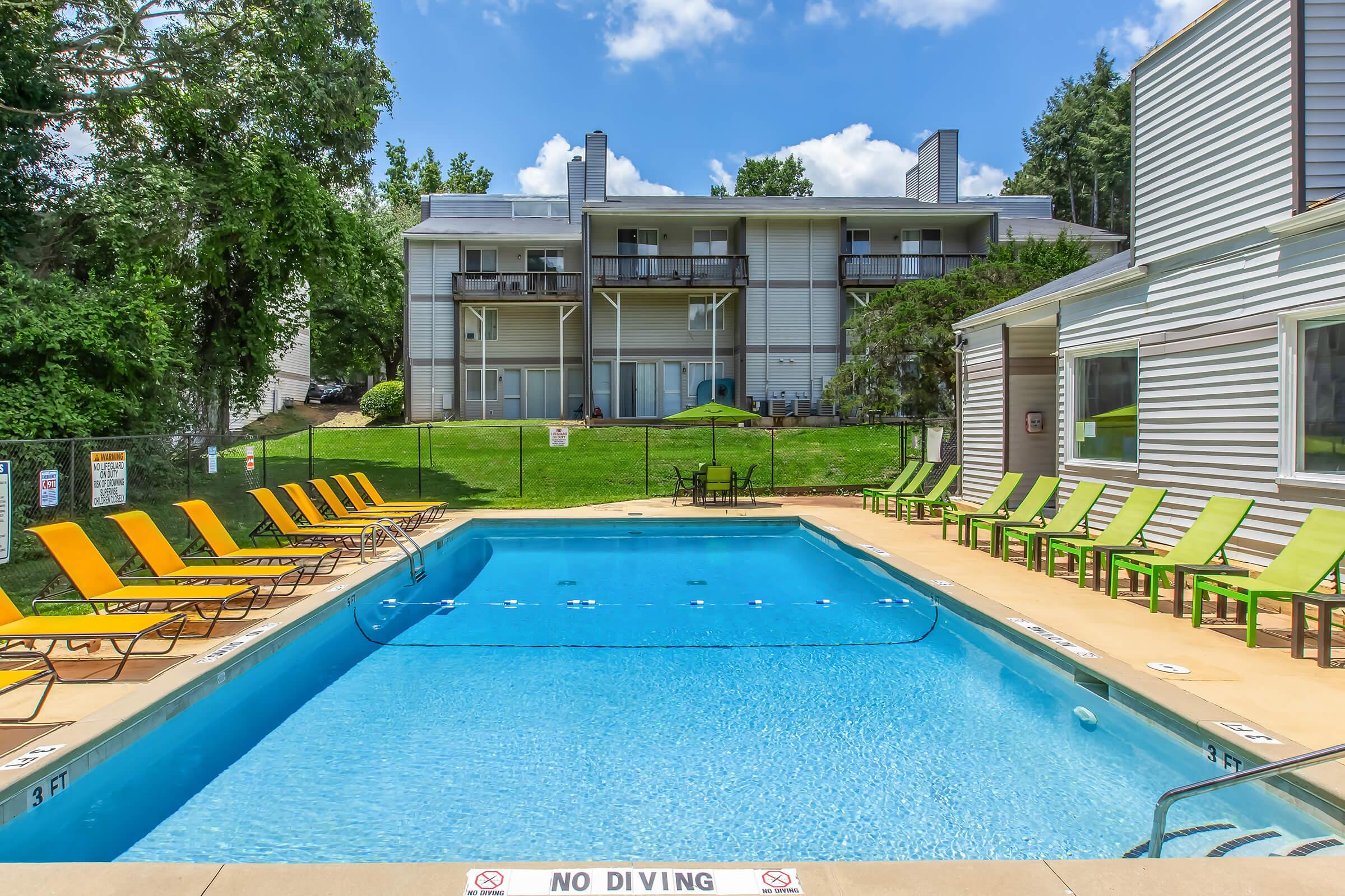 A clear blue swimming pool surrounded by lounge chairs in yellow and green. In the background, there are several multi-story residential buildings set among trees under a bright blue sky with some clouds. A "No Diving" sign is visible at the pool's edge.