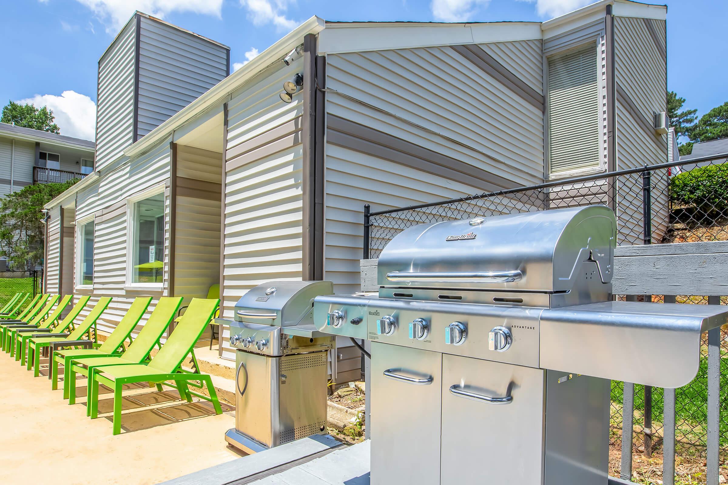 A modern outdoor space featuring two silver grills placed beside vibrant green lounge chairs. The background includes a light-colored building with multiple stories and large windows, set against a clear blue sky. The area is well-maintained and inviting, perfect for outdoor gatherings.