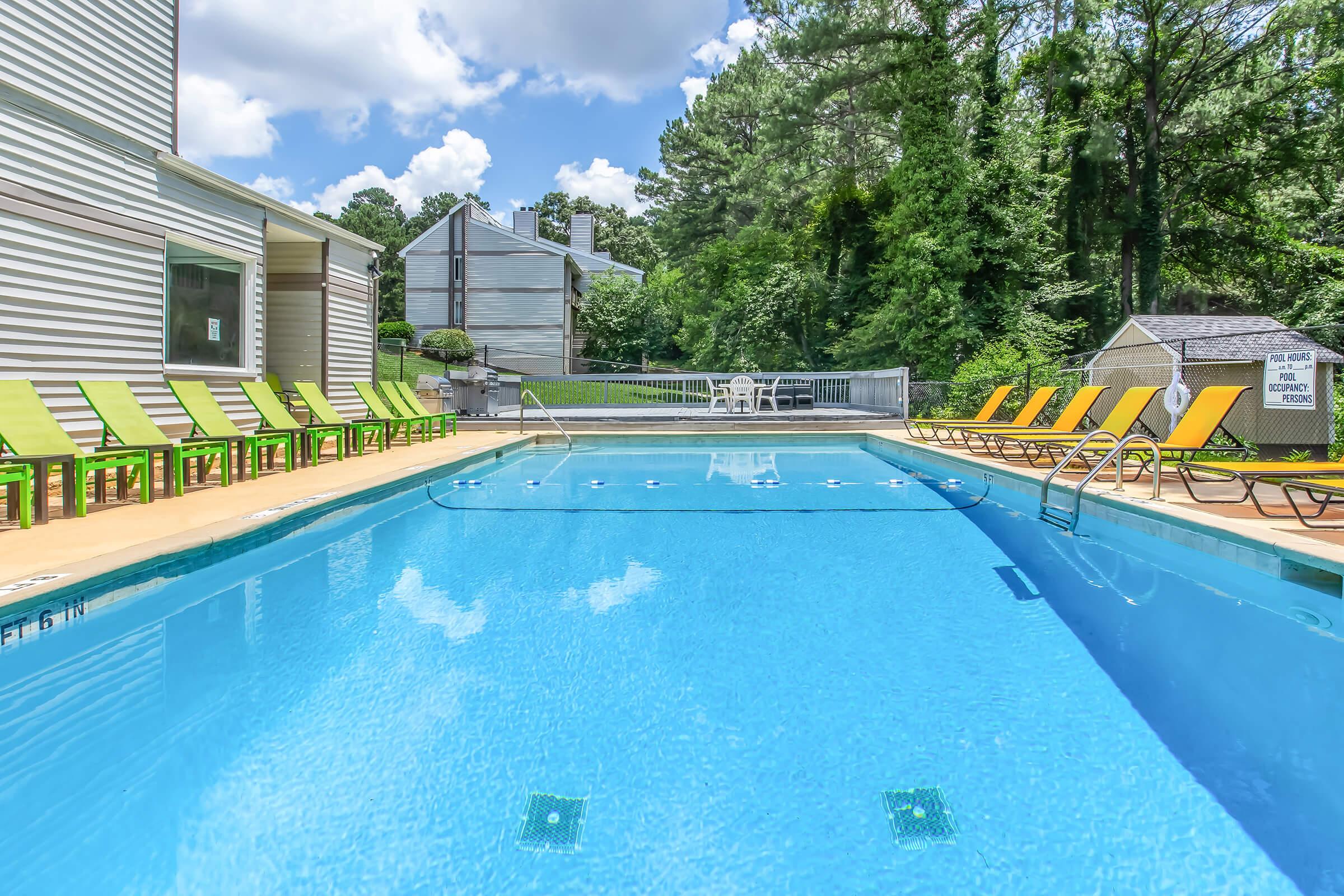 A clear blue swimming pool surrounded by green lounge chairs on one side. Lush greenery and trees are visible in the background, alongside a few residential buildings. A small seating area with white chairs is situated near the pool, under a bright blue sky with some white clouds.