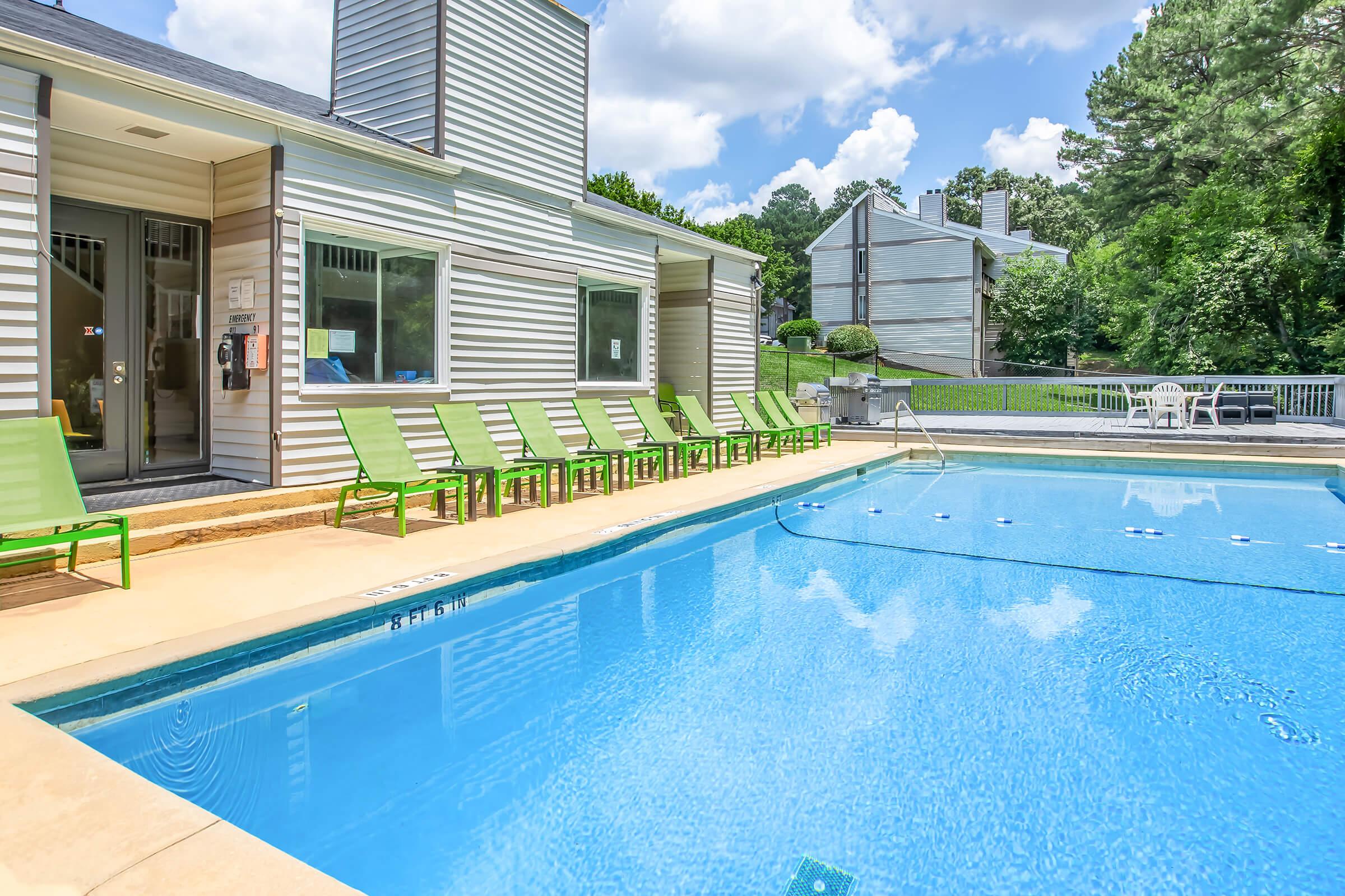 A view of a sparkling blue swimming pool surrounded by green lounge chairs. In the background, there are several residential buildings and lush greenery under a partly cloudy sky. The pool area features clean, inviting water, perfect for relaxation and recreational activities.