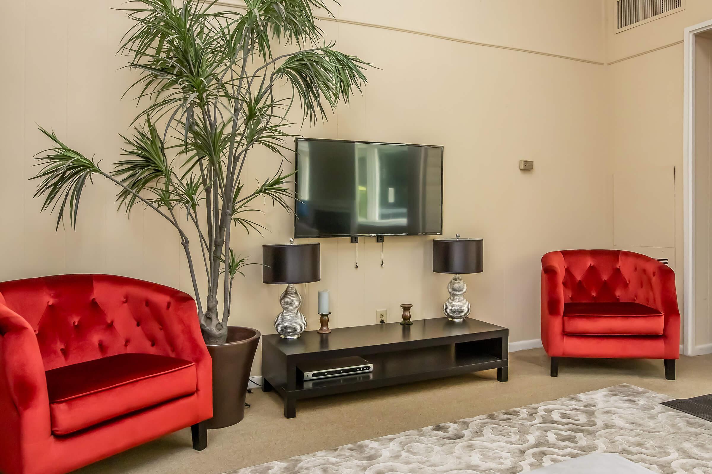 A cozy living room featuring two red tufted chairs, a dark wooden TV stand with a television, and decorative lamps. A potted plant adds a touch of greenery, while a light-colored rug enhances the inviting atmosphere. The walls are painted a soft beige, creating a warm and welcoming space.