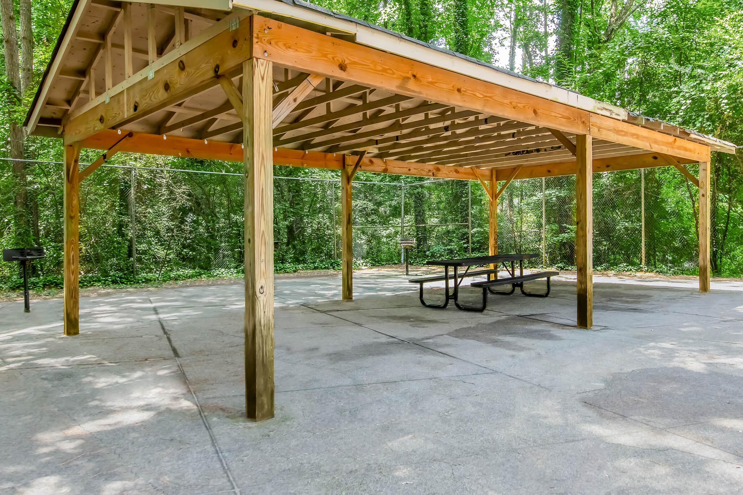 A wooden pavilion with a gabled roof, featuring wooden beams and a concrete floor. Underneath, there is a picnic table, and the surrounding area is shaded by trees. The setting is peaceful, ideal for gatherings or outdoor activities.