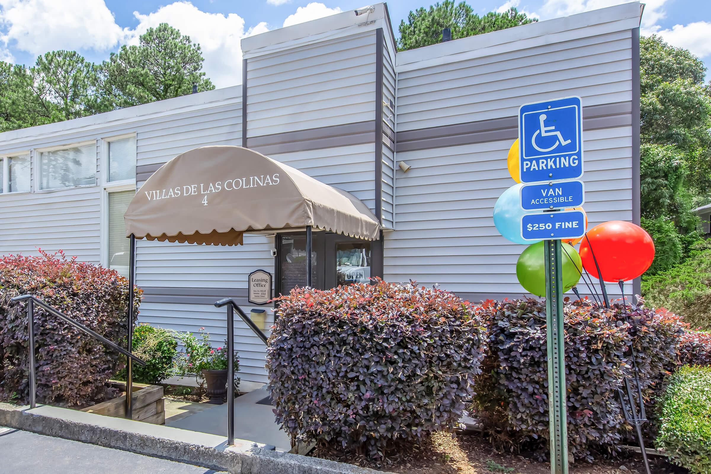 Exterior view of a building labeled "Villas de las Colinas" with a brown awning. There are colorful balloons next to a parking sign indicating a handicapped parking spot with a $250 fine for violations. Surrounding greenery includes bushes and the sky is partly cloudy.