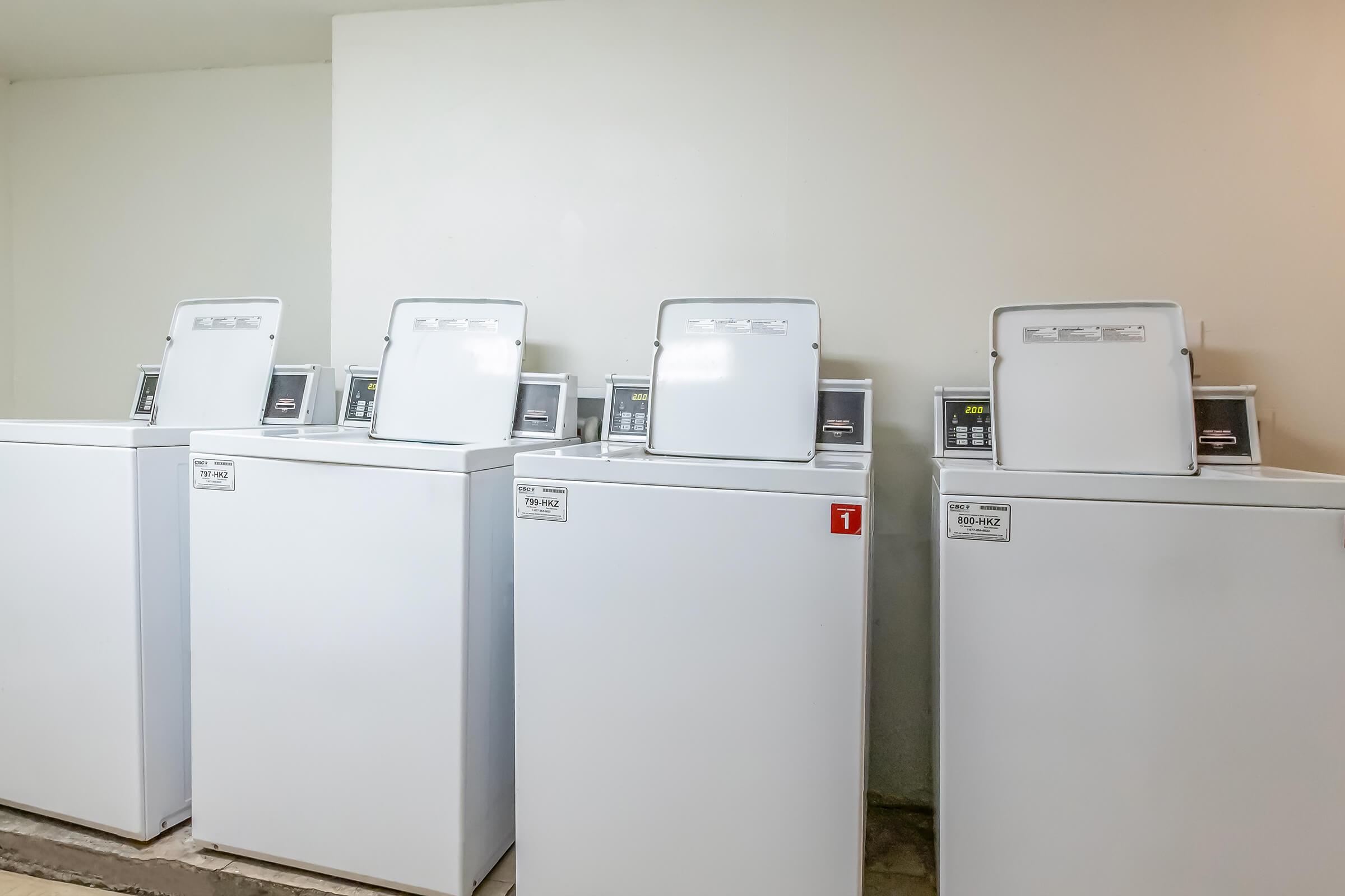 A row of top-loading washing machines in a laundry room setting. The machines are white with digital displays and are positioned on a concrete floor against a plain wall, conveying a clean and utilitarian atmosphere.