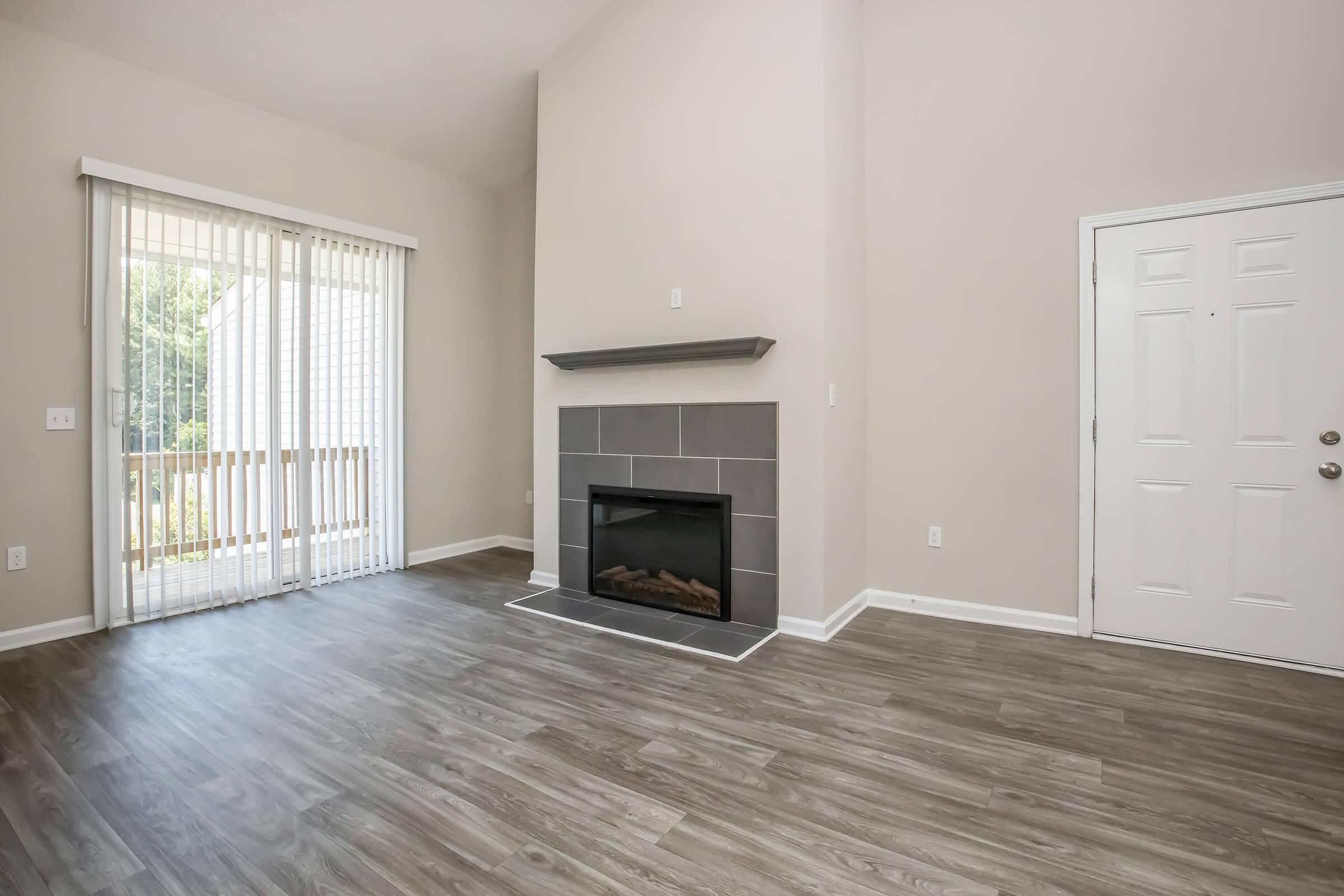 A spacious living room featuring light-colored walls, a modern fireplace with a gray mantel, and large sliding glass doors leading to a balcony. The flooring is a light wood style, and a front door is visible on the right. Natural light fills the room, creating a welcoming atmosphere.