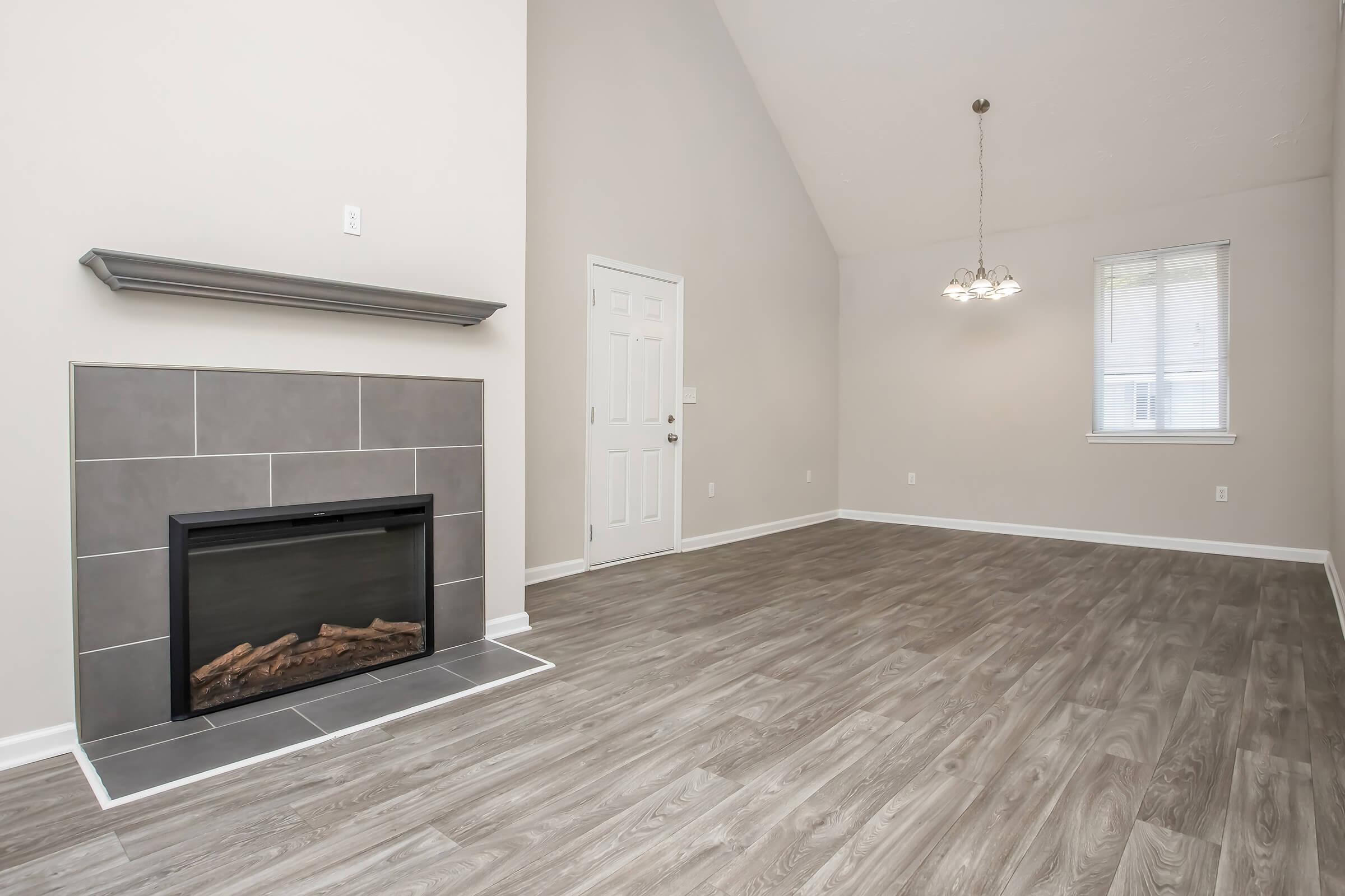 A spacious living room featuring a high ceiling, light beige walls, and gray wood-like flooring. A modern fireplace with a tiled surround is positioned against one wall. Natural light enters through a window, and there is a door leading outside. A chandelier hangs from the ceiling, adding a decorative touch.