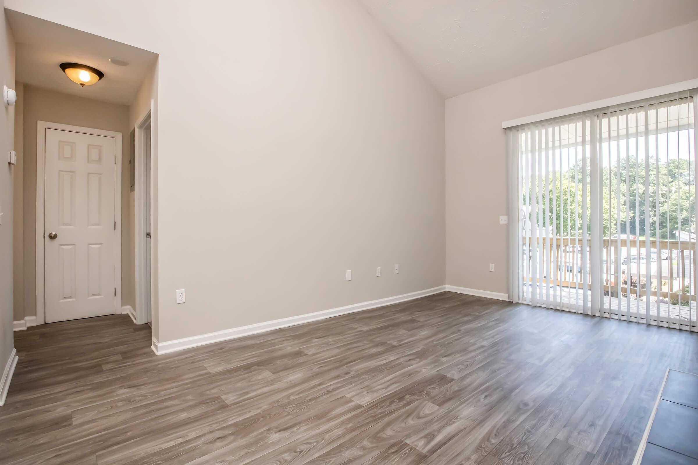 A spacious, empty living room featuring light-colored walls and a high ceiling. The floor has a wood-like finish, and there is a large set of sliding glass doors leading to an outdoor balcony. A hallway with a door is visible on the left side. Bright natural light fills the room.