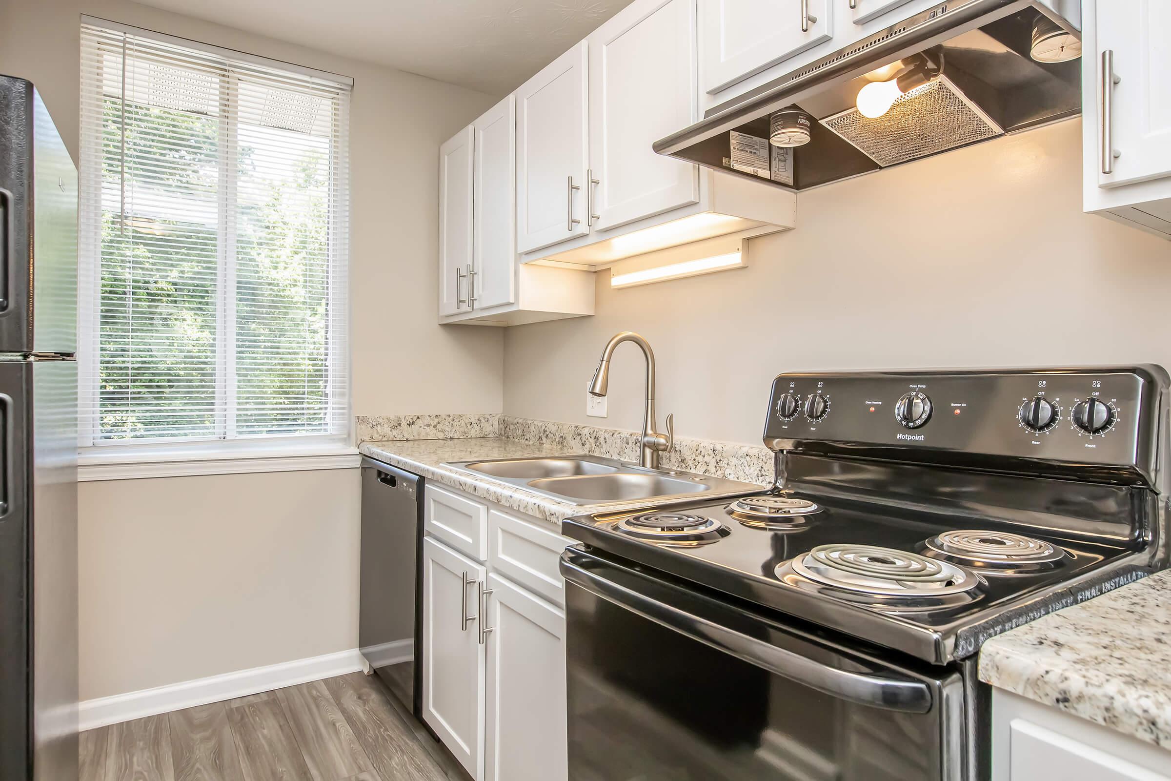 Modern kitchen featuring white cabinetry, a black stove with four burners, and granite countertops. A stainless steel sink is positioned between the stove and a dishwasher. Natural light floods the space through a window, illuminating the sleek design and functional layout.