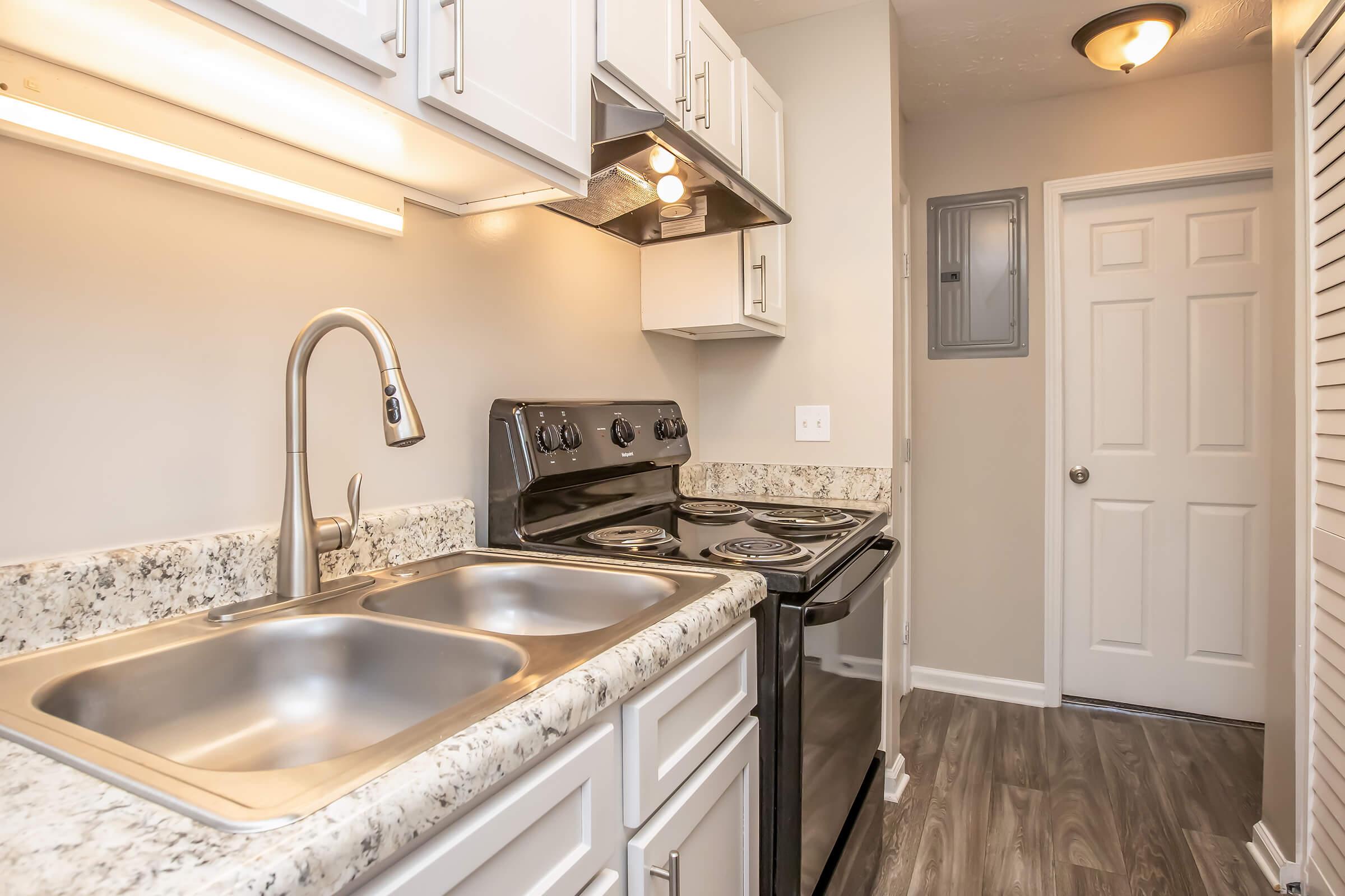 A modern kitchen featuring granite countertops, a stainless steel double sink, an electric stove, and white cabinetry. The kitchen is well-lit with under-cabinet lighting and has a doorway leading to another room, showcasing a neutral color palette and contemporary finishes.