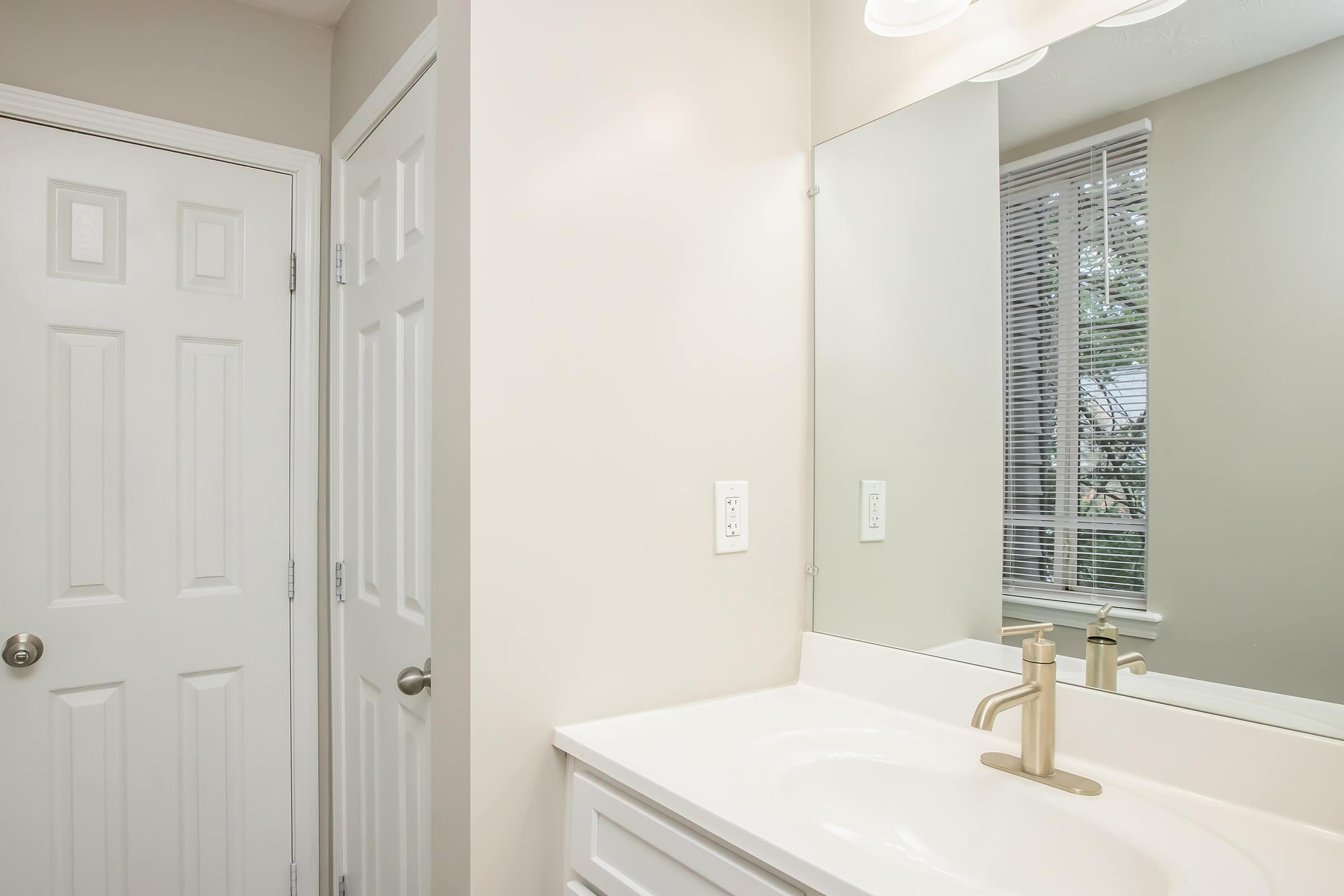 A clean, neutral-toned bathroom featuring a white vanity with a modern faucet, a rectangular mirror above the sink, and two doors visible. Natural light streams through a window with blinds, illuminating the space. The walls are painted in a light beige color, enhancing the bright and airy feel.