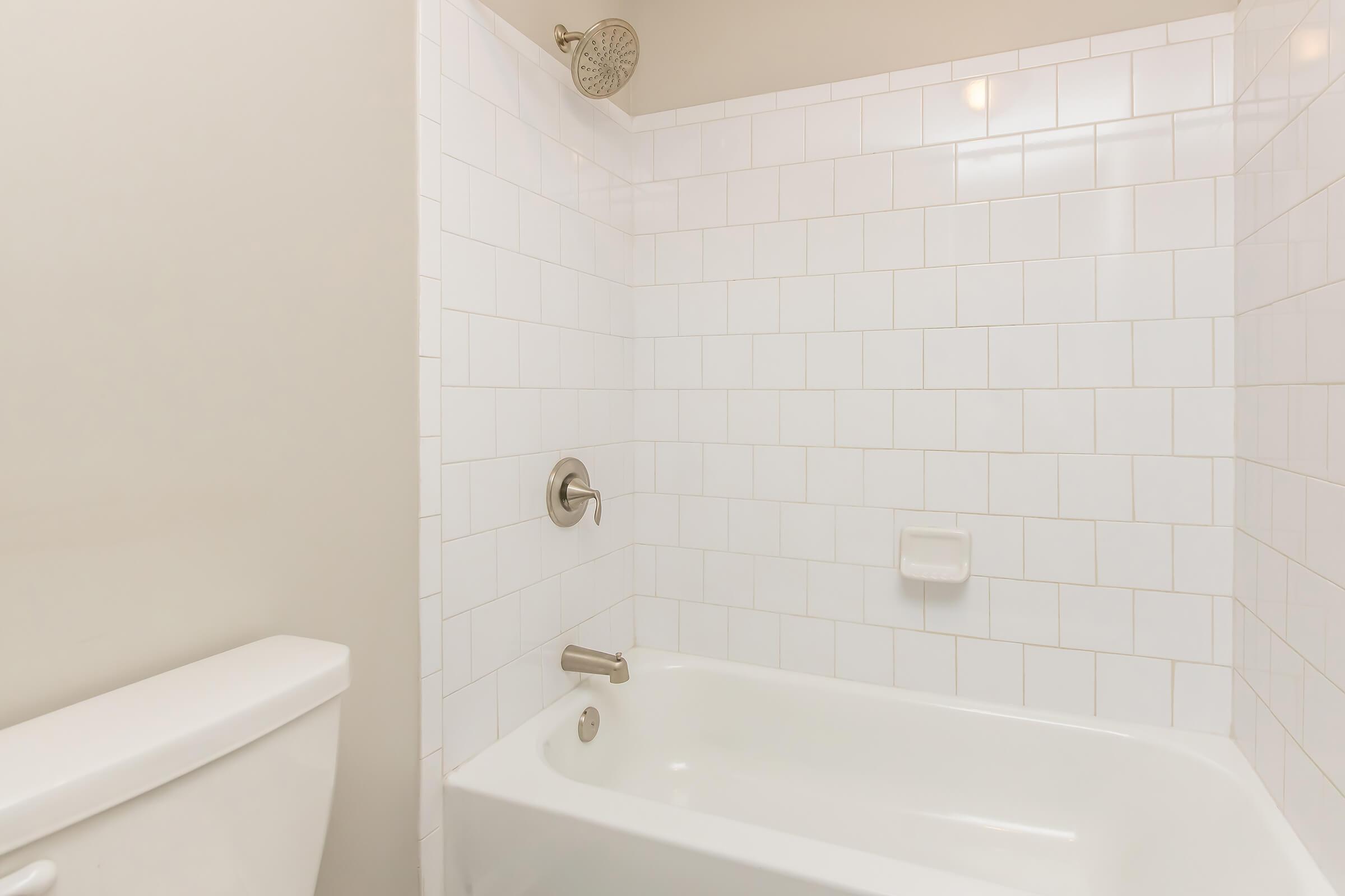 A clean, minimalistic bathroom featuring a white tiled shower area with a bathtub, a showerhead, and a chrome faucet. The toilet is in the corner, and the walls are painted a soft color, creating a bright and airy feel.