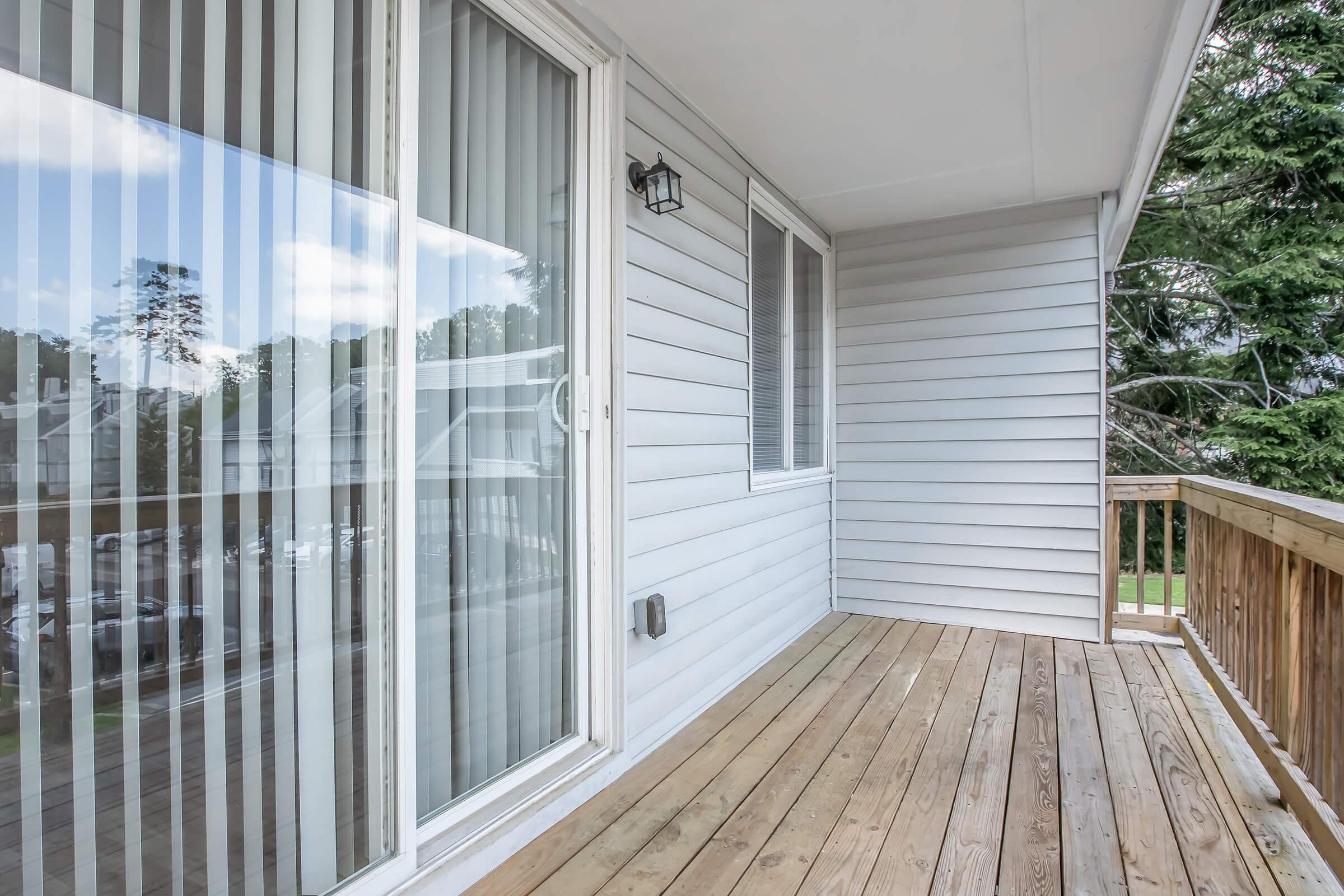 A spacious balcony featuring wooden flooring and large glass sliding doors with vertical blinds. The exterior walls are clad in light-colored siding, and a small light fixture is visible on the wall. Green trees can be seen in the background, enhancing the peaceful atmosphere of the space.