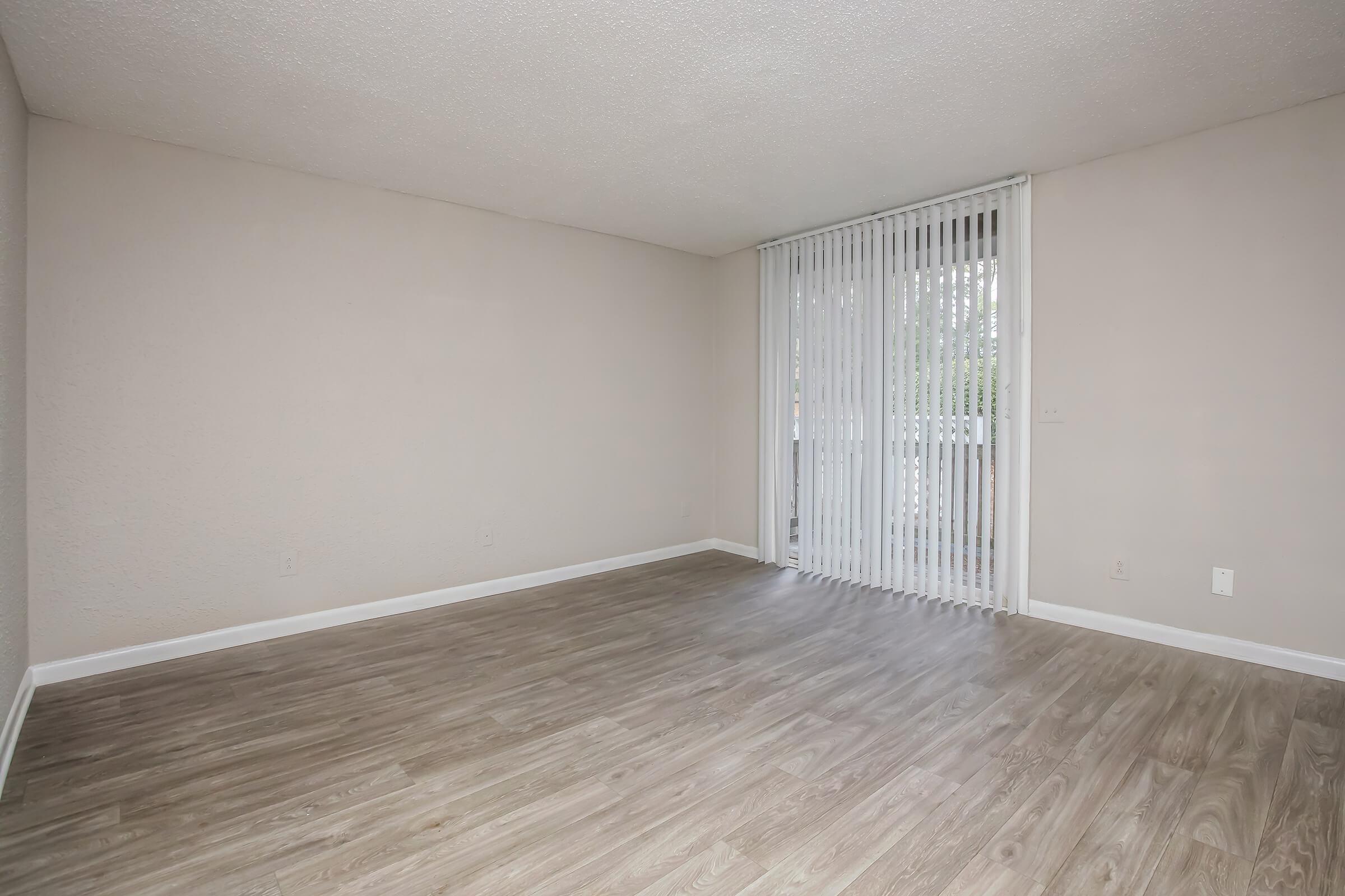 Empty, well-lit room with light-colored walls and a large window covered by vertical blinds. The flooring is modern, resembling wood. There is no furniture or décor, giving a spacious and minimalistic appearance.