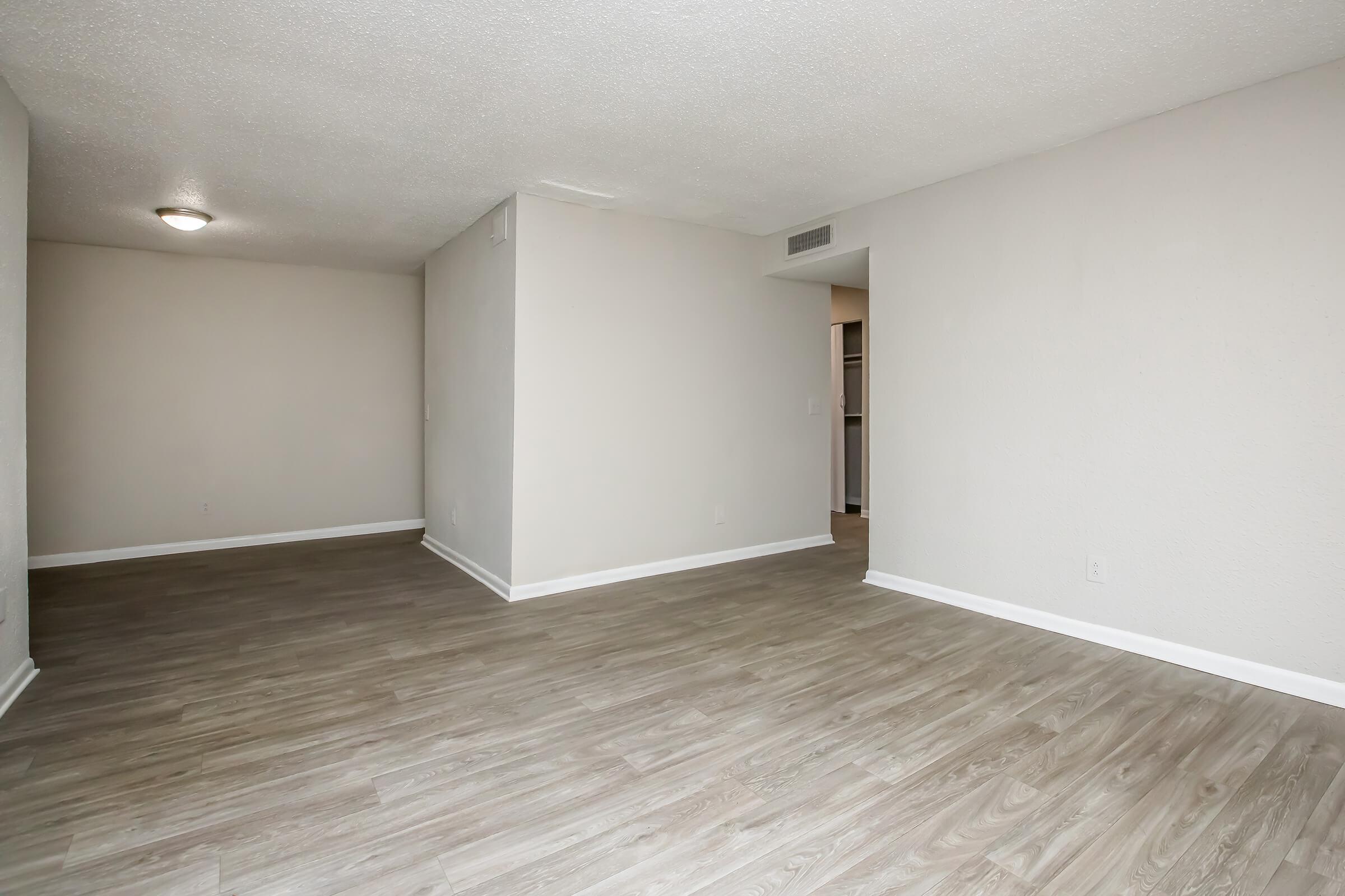 A spacious, empty living room featuring light-colored walls and a smooth, laminate floor. Natural light fills the area, enhancing the sense of openness. A doorway leads to another room, possibly a kitchen or closet, suggesting a modern layout. The overall atmosphere is clean and minimalistic.
