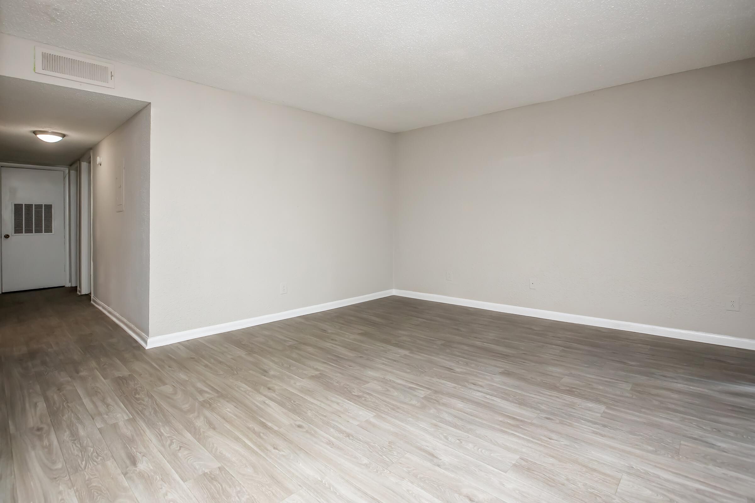 Empty room with light-colored walls and hardwood flooring. A doorway is visible on the left side leading to a hallway. The space is well-lit by overhead lighting, providing a bright and open atmosphere. The absence of furniture emphasizes the spaciousness of the area.