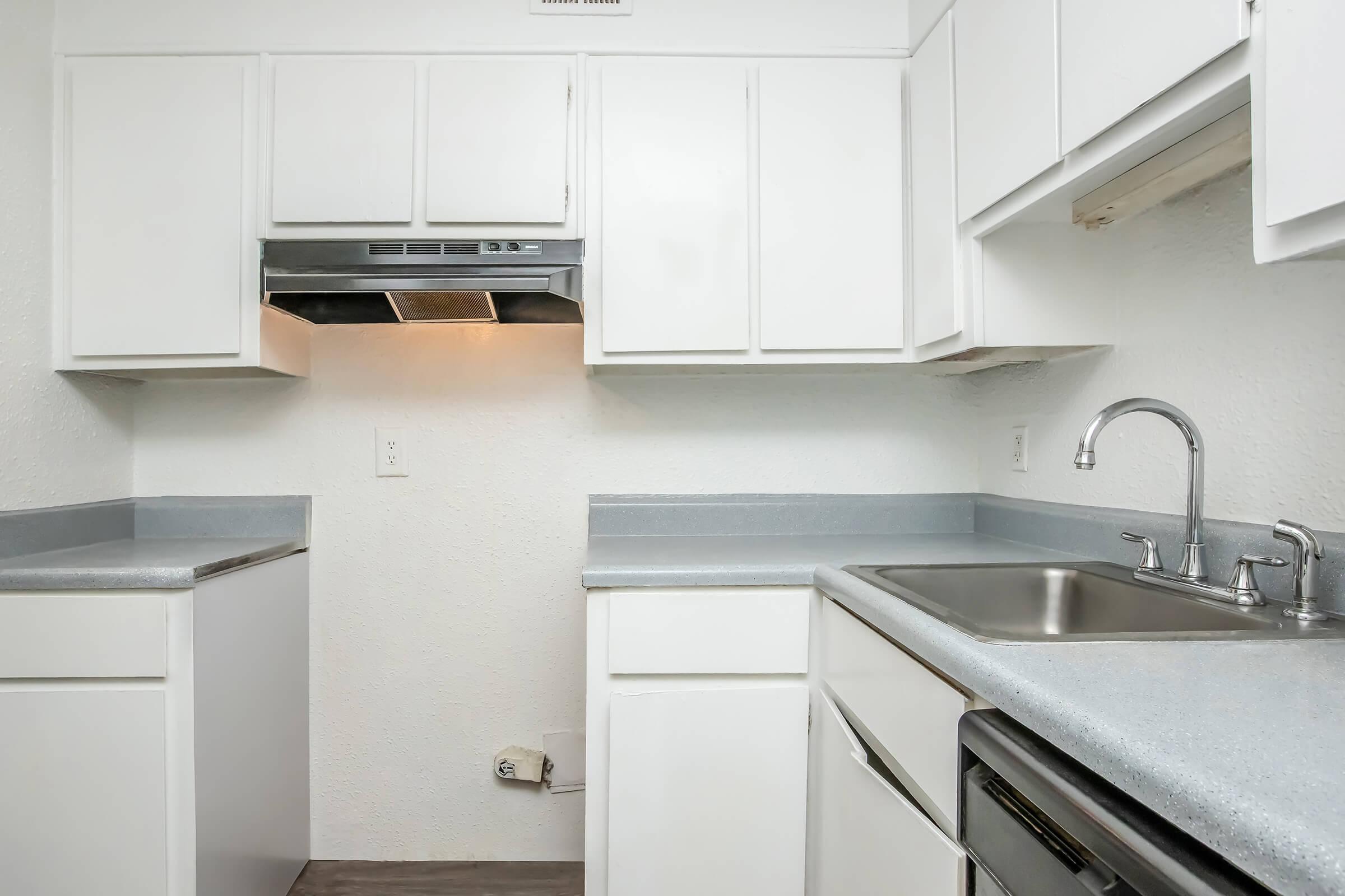 A clean, modern kitchen featuring white cabinetry, a gray countertop, a stainless steel sink, and a stove. The kitchen includes an under-cabinet range hood and has a minimalist design with ample space for cooking. The walls are painted white, creating a bright and airy atmosphere.