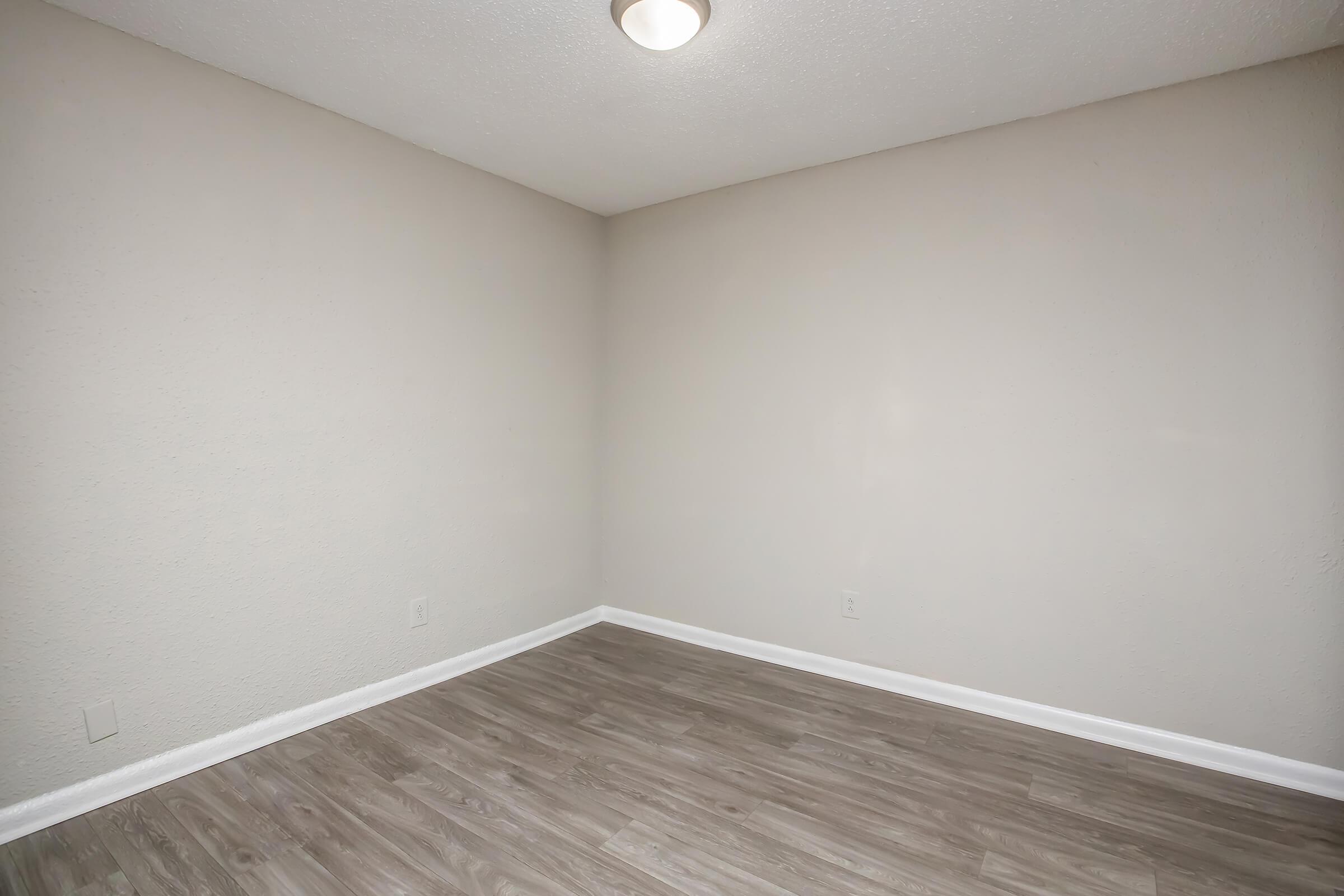 A small empty room with light gray walls and a light wood floor. The ceiling has a circular light fixture. There are no furniture or decorations present, creating a minimalist and spacious feel.