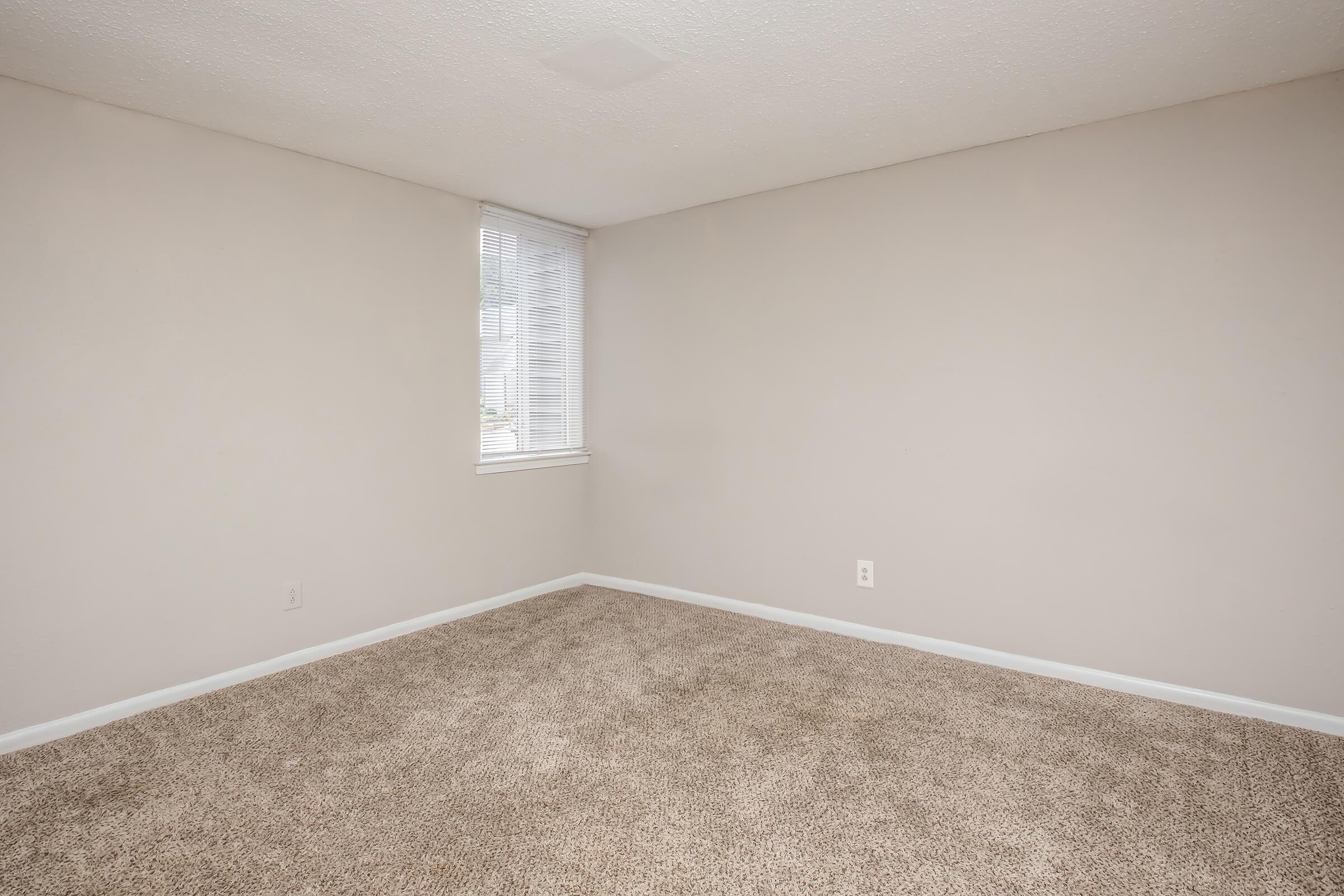 A small, empty room with light beige walls and a window with blinds. The floor is covered in light brown carpet, and there are no furniture or decorations, creating a spacious and minimalistic atmosphere.