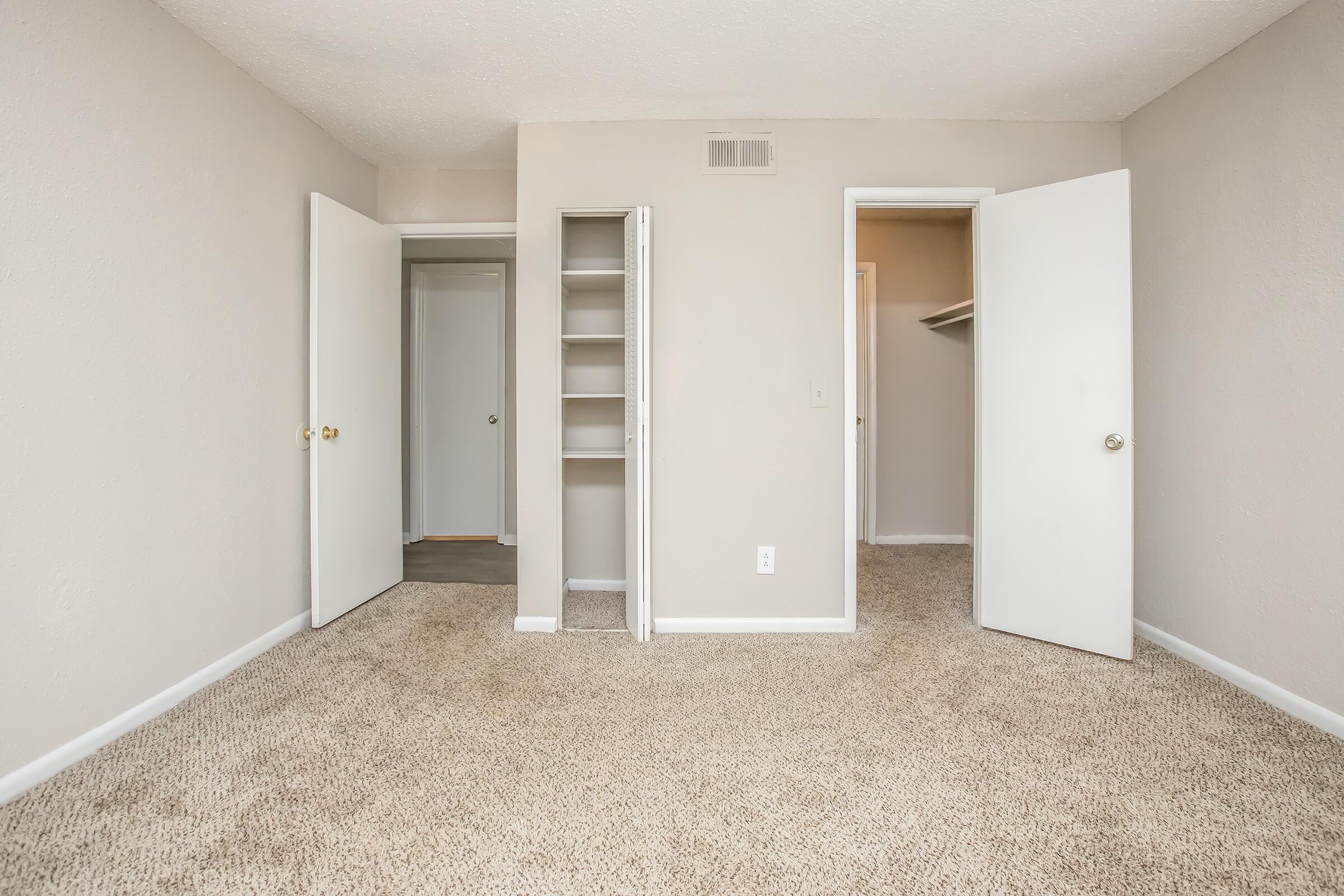 A vacant room with beige walls and carpet, featuring two open doors leading to closets. One door is on the left with visible shelves, and the other door on the right leads to a closet space. The room is well-lit with natural light creating a spacious feel.
