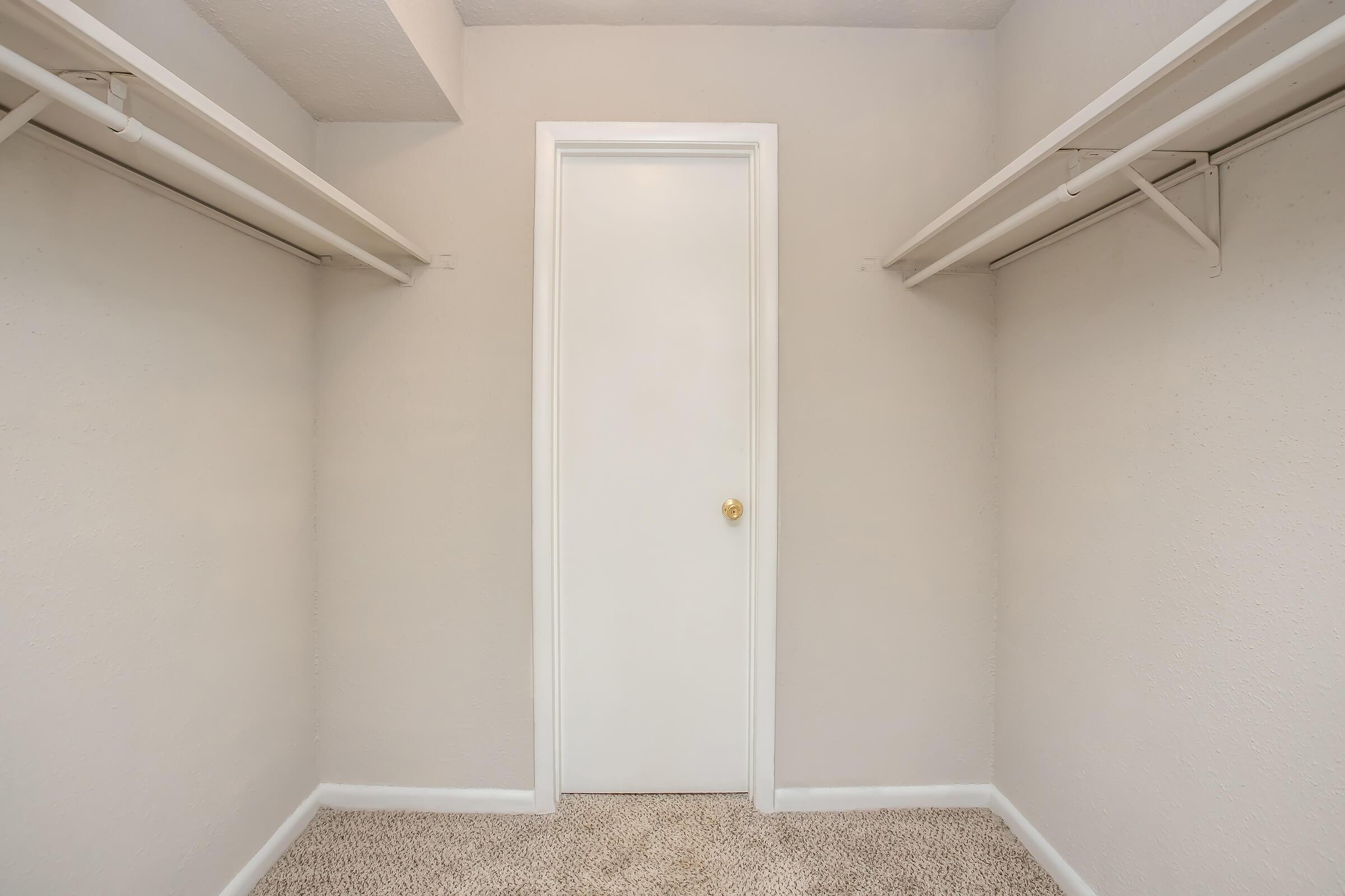 Empty closet with two metal shelves on either side and a closed white door in the center. The walls are painted light beige, and the carpet is a neutral color, creating a simple, minimalistic space.