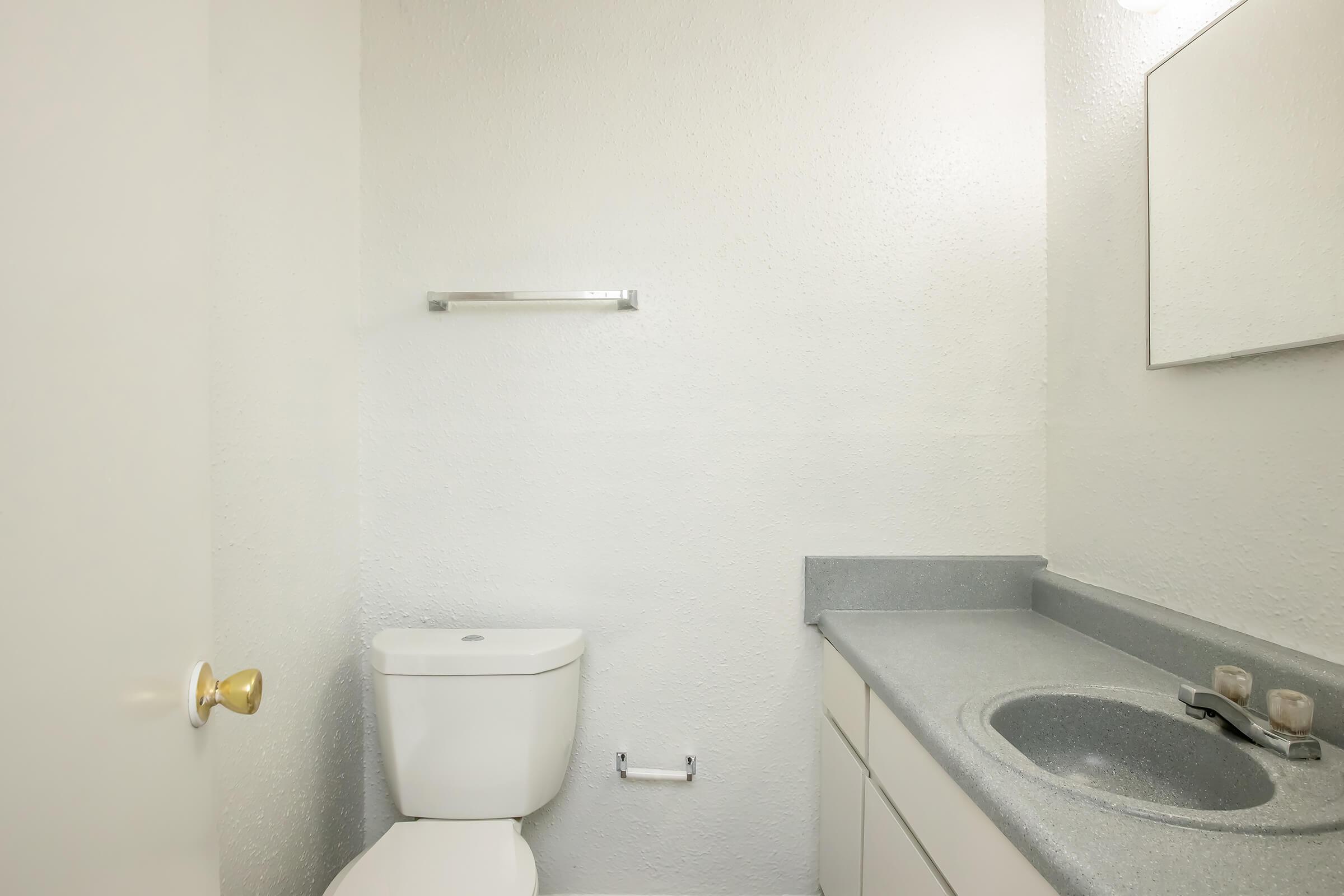 A clean, modern bathroom featuring a white toilet, a gray countertop with a sink, and a mirror. The walls are painted white, and there is a towel rack mounted on the wall. The space is well-lit and minimalistic, emphasizing a simple aesthetic.
