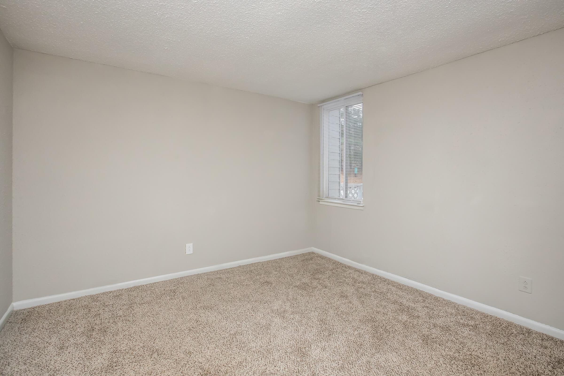 A vacant room with light beige walls and a plush carpet. There is a small window with blinds, allowing natural light to enter. The space is empty, making it suitable for various uses like a bedroom or office, and has a clean, neutral aesthetic.