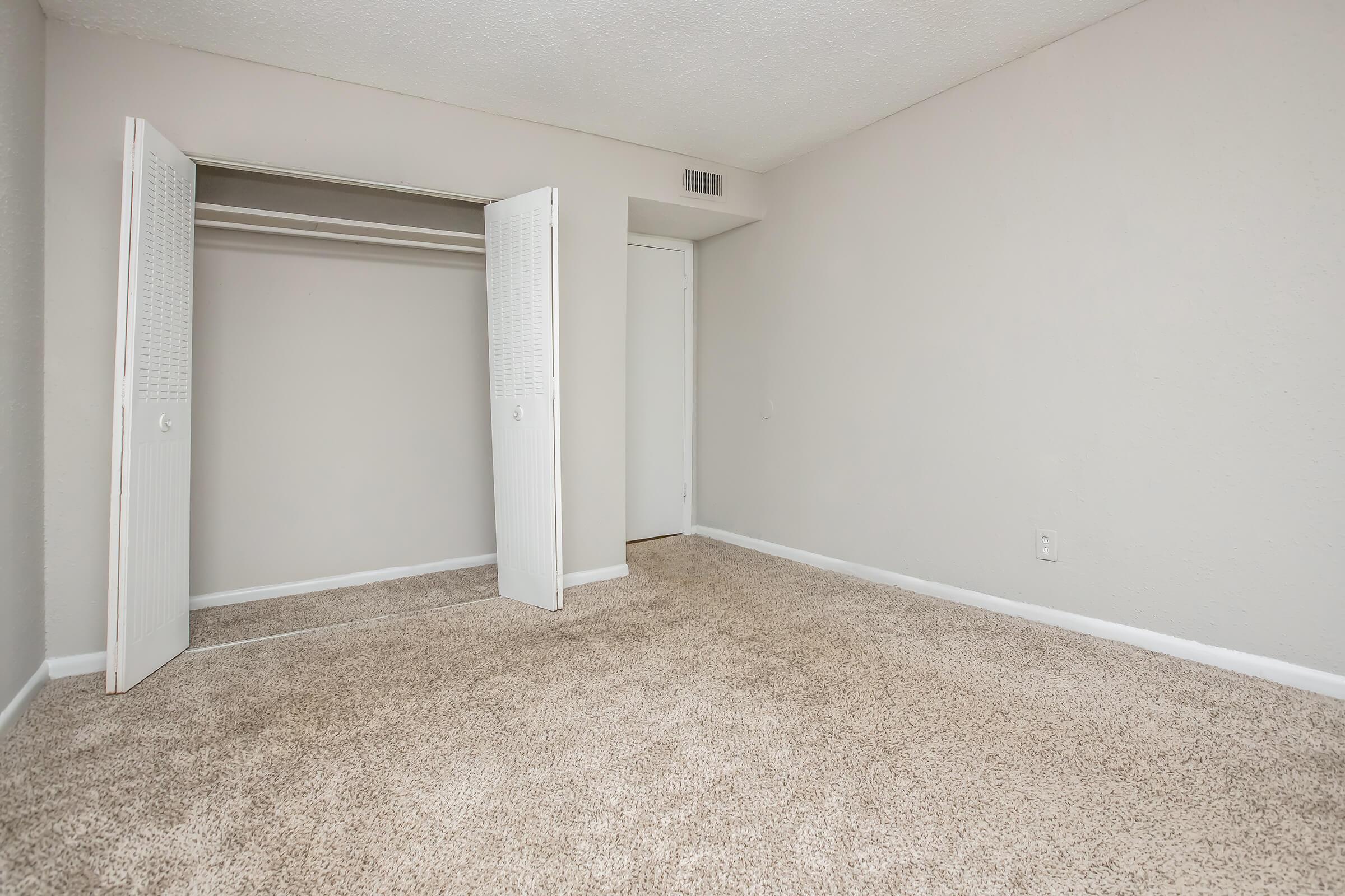 An empty room with light beige walls and a carpeted floor. There is a closed door on the right and an open closet with white doors on the left, providing a view of the space inside. The room is well-lit and lacks furniture, creating a minimalistic atmosphere.
